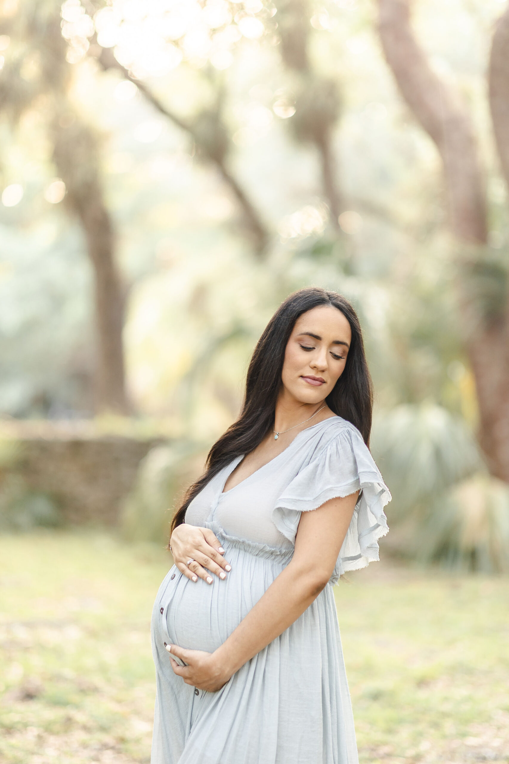 A mother to be stands in a park at sunset holding her bump and gazing down her shoulder in a flowing maternity gown after exploring baby shower venues in coral gables