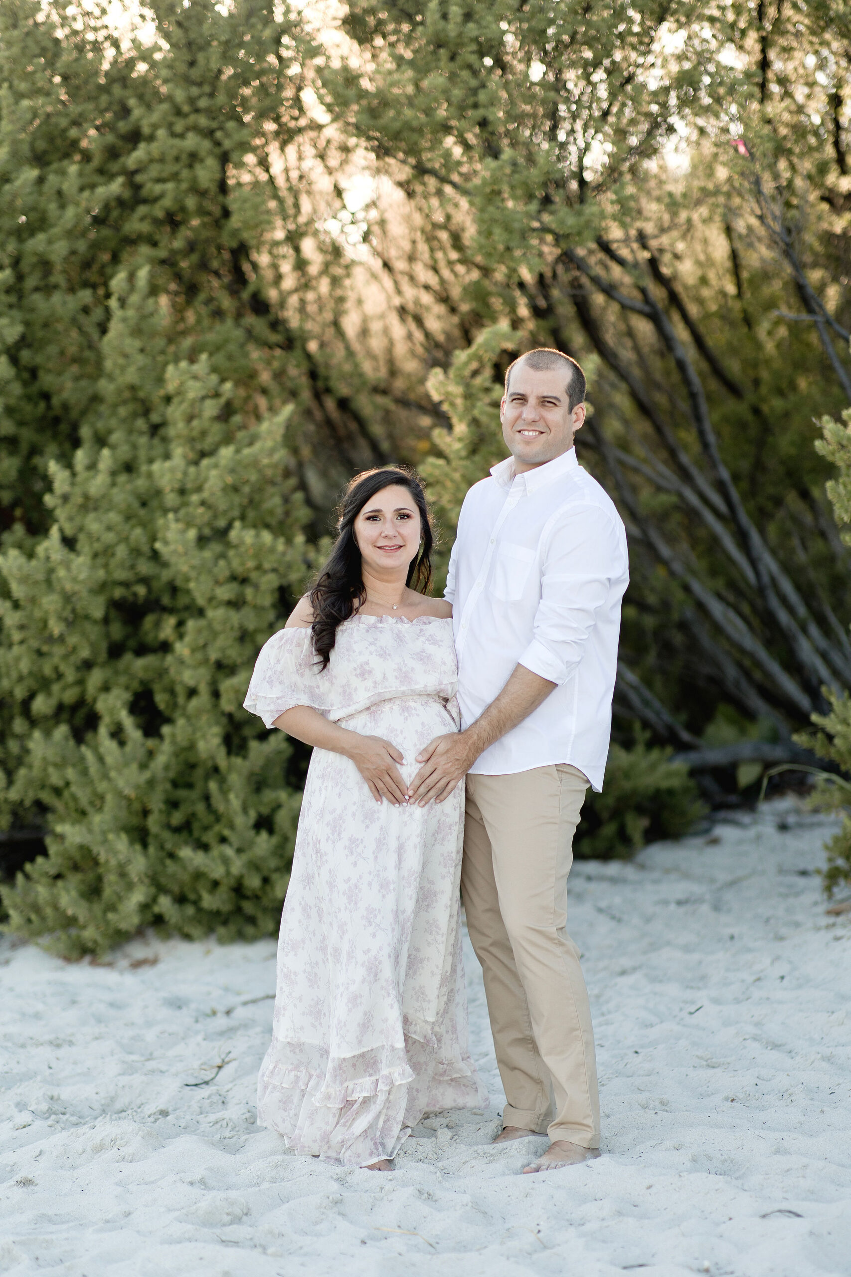 A happy pregnant couple stand with hands on the bump on a beach dune at sunset