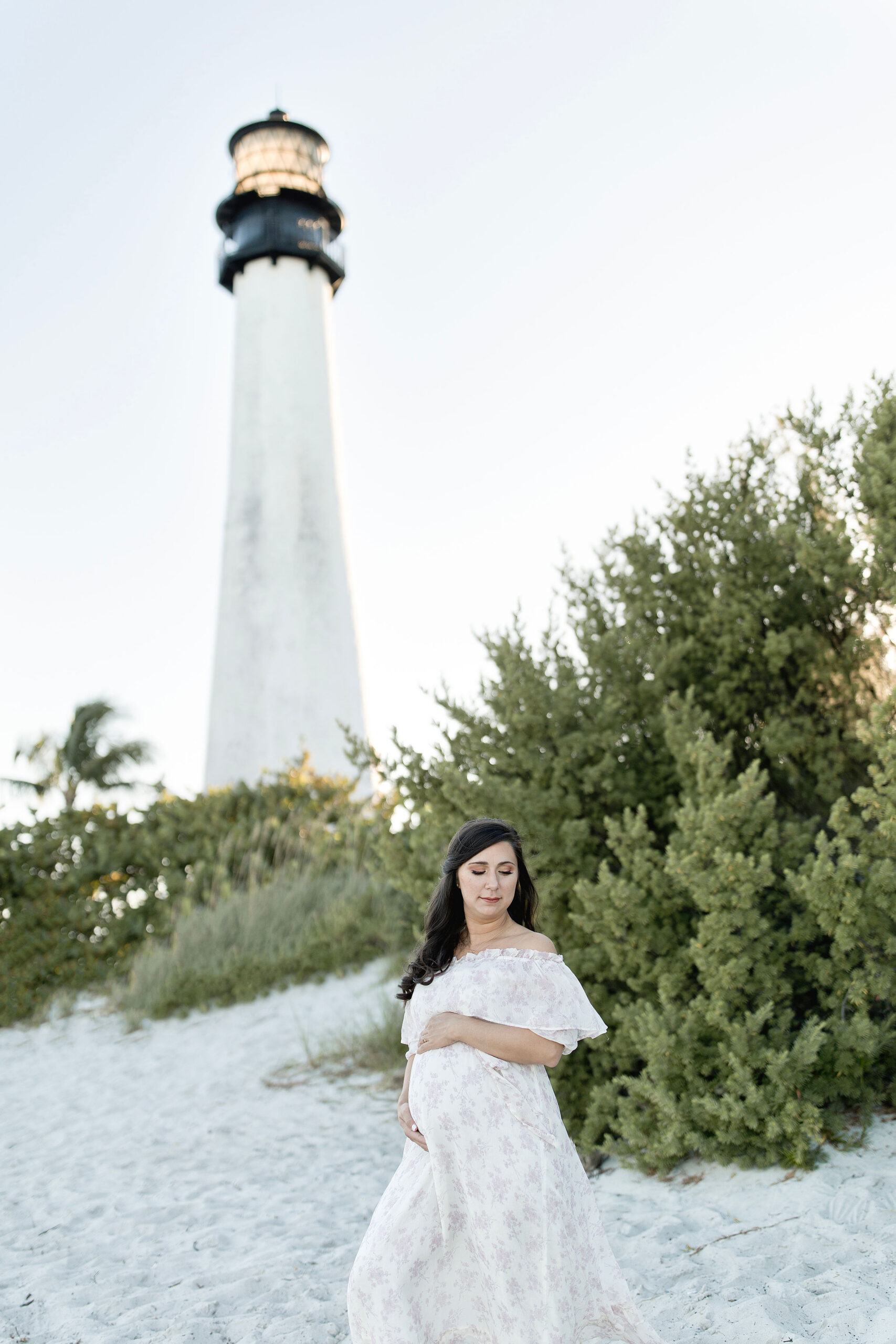 A mother to be stands on a beach under a lighthouse with hands on bump in a floral print dress after meeting obgyns in coral gables