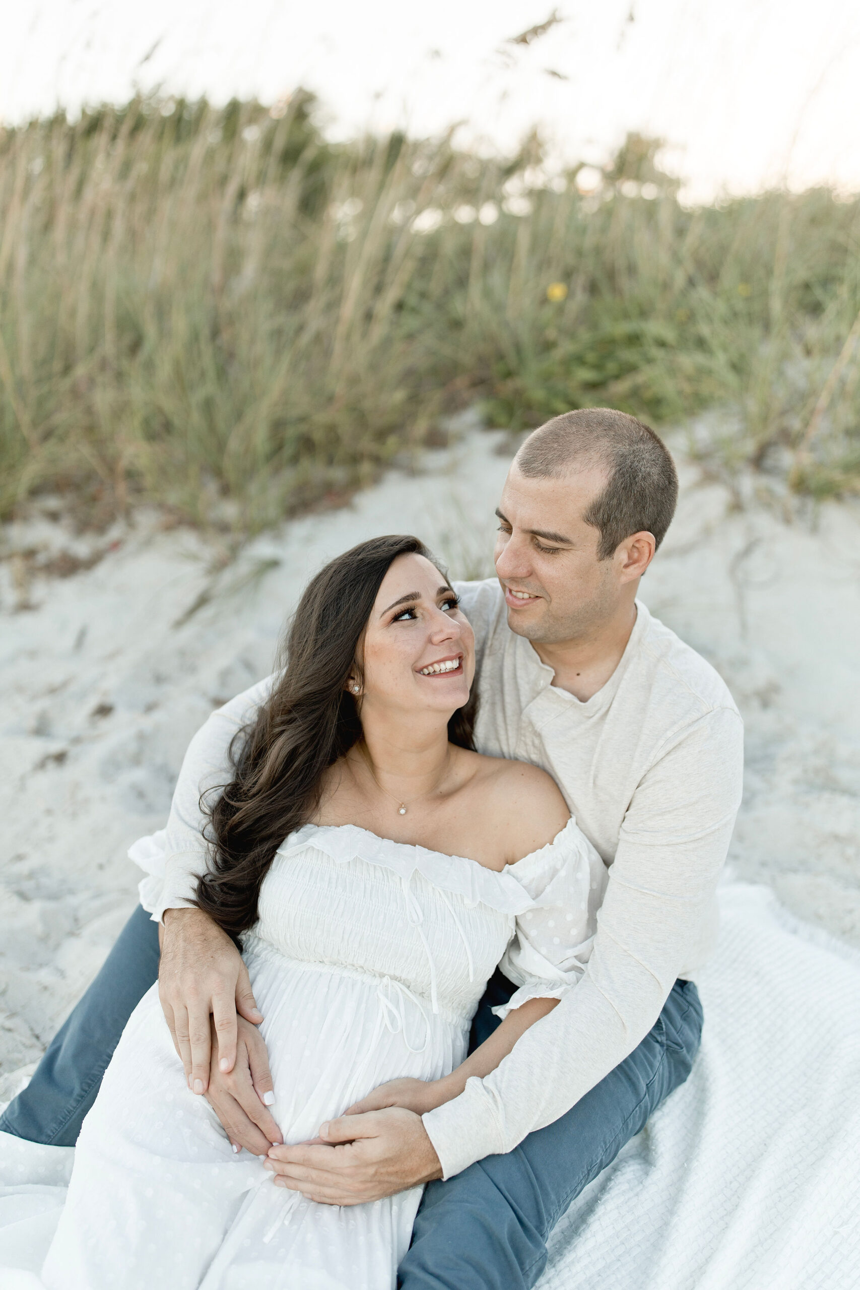 A happy pregnant woman smiles while leaning into the lap of her husband on a beach after finding obgyns in coral gables