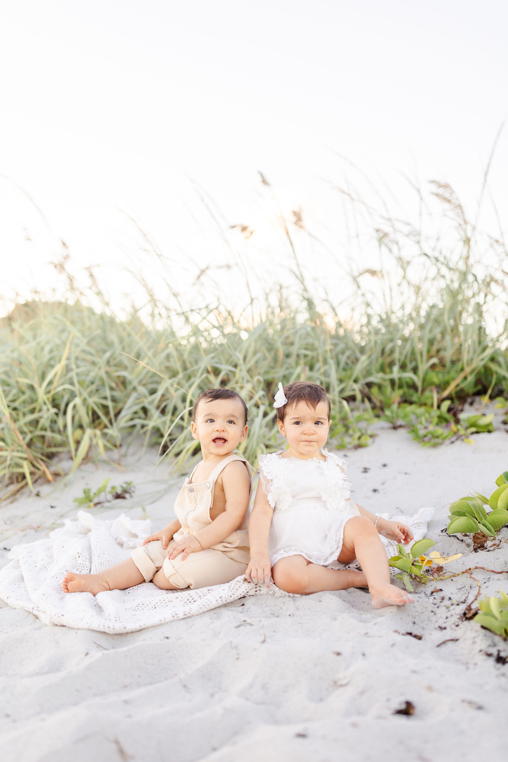 Twin toddlers sit on the beach at sunset near the dune grass in white and cream