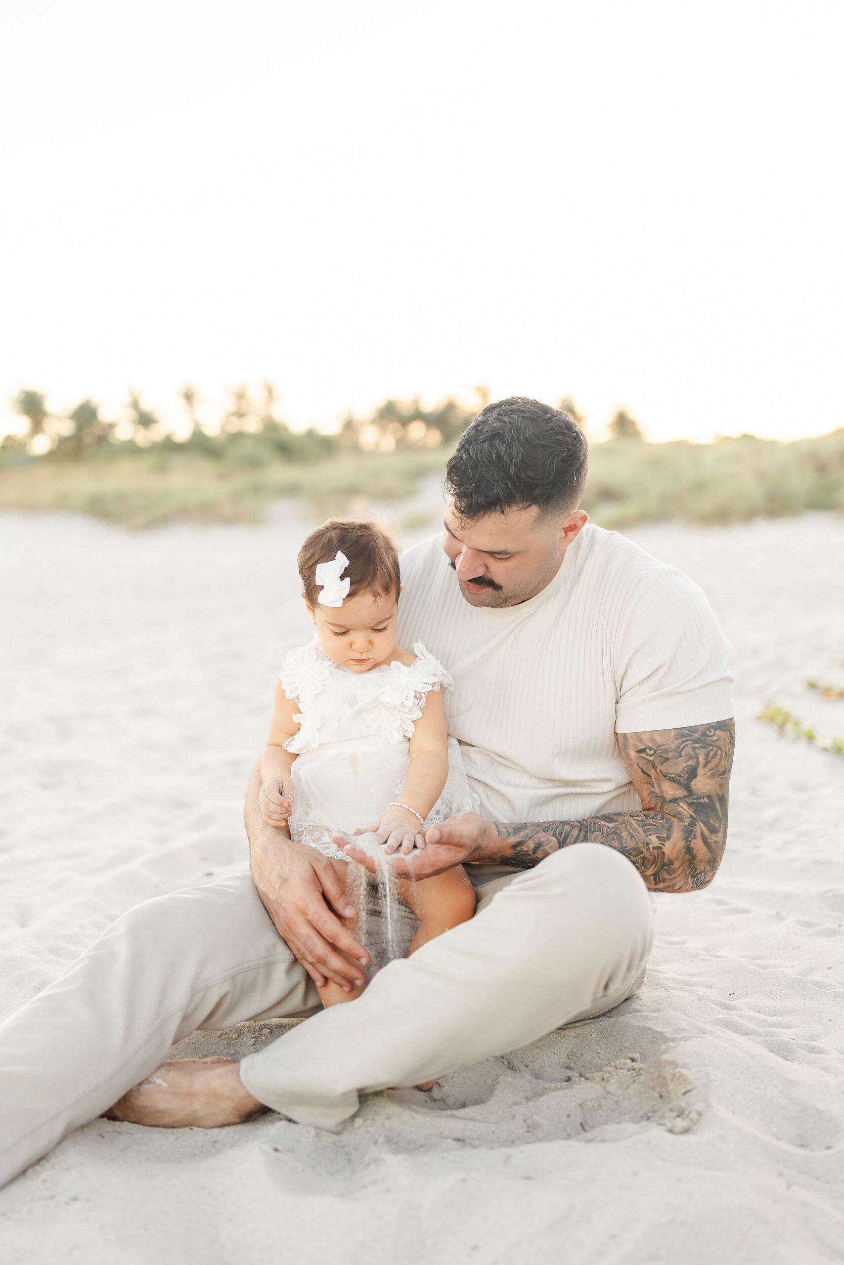 A toddler girl in a white dress plays with sand on the beach sitting in dad's lap at sunset after some swim lessons in coral gables