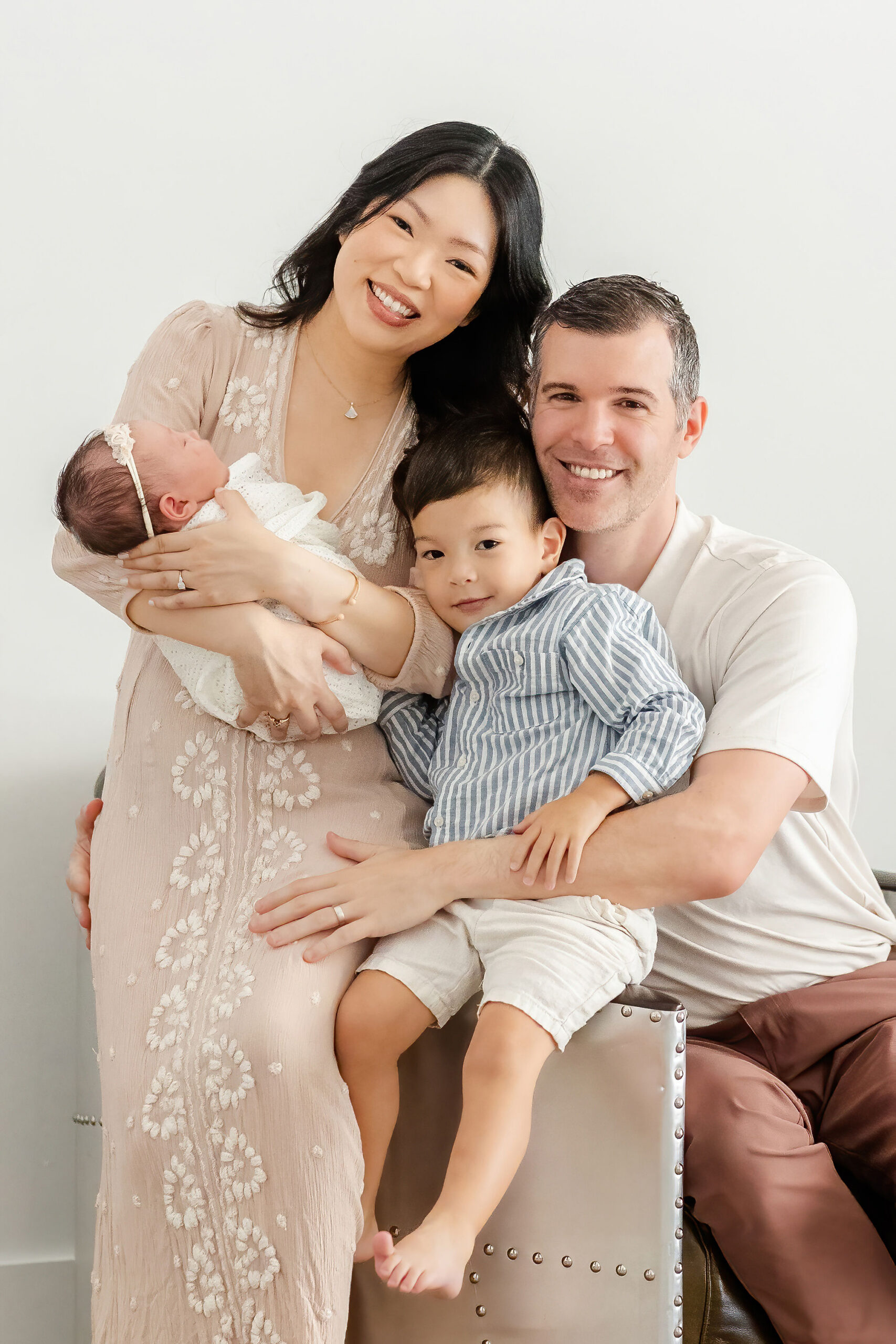 Happy mom and dad sit on a modern chair holding a sleeping newborn girl and toddler son sitting on the arm