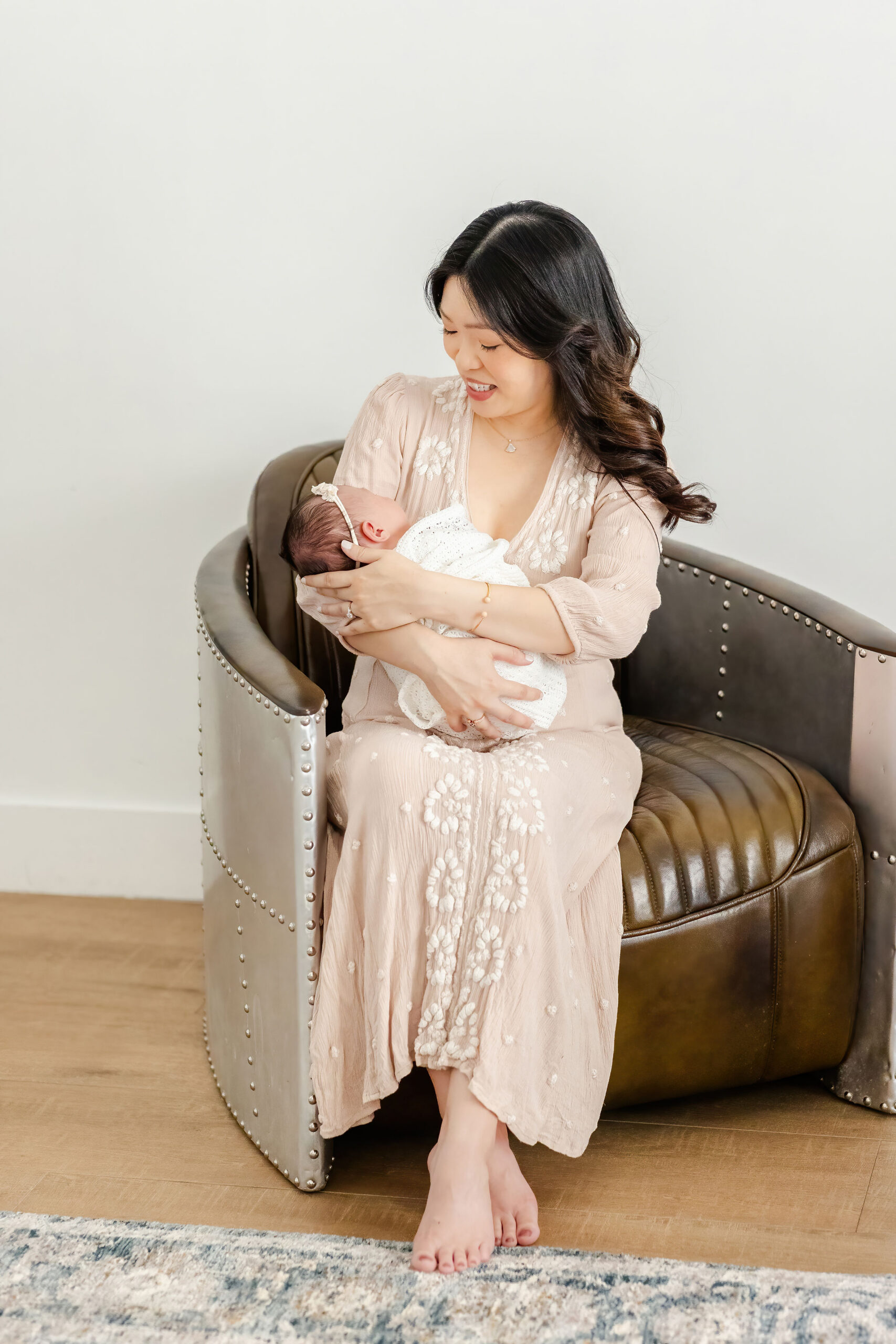 A new mom sits on a leather chair cradling her sleeping newborn daughter in a white swaddle after visiting the wander shop
