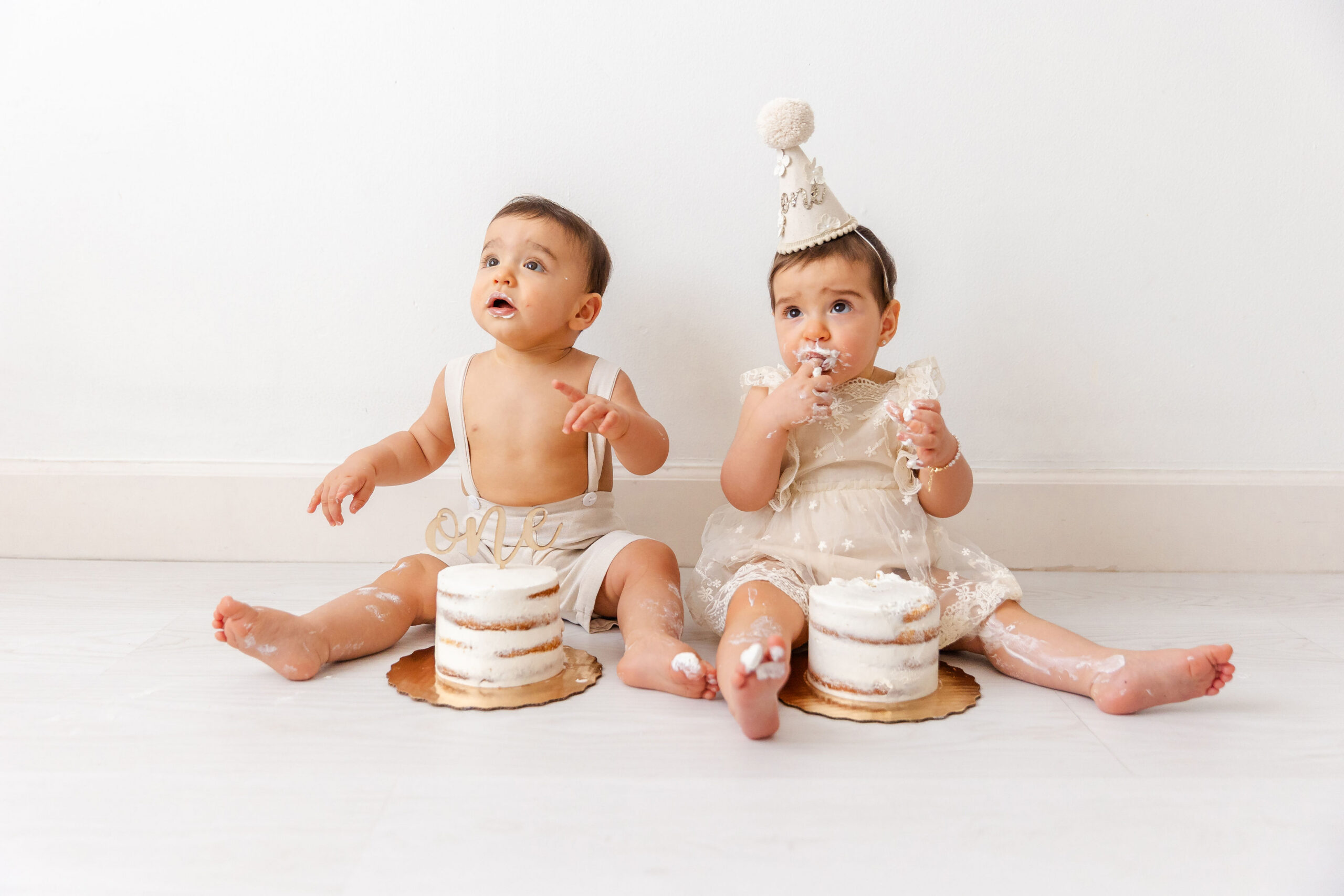 Twin brother and sister sit on the floor behind matching birthday cakes in cream outfits covered in icing