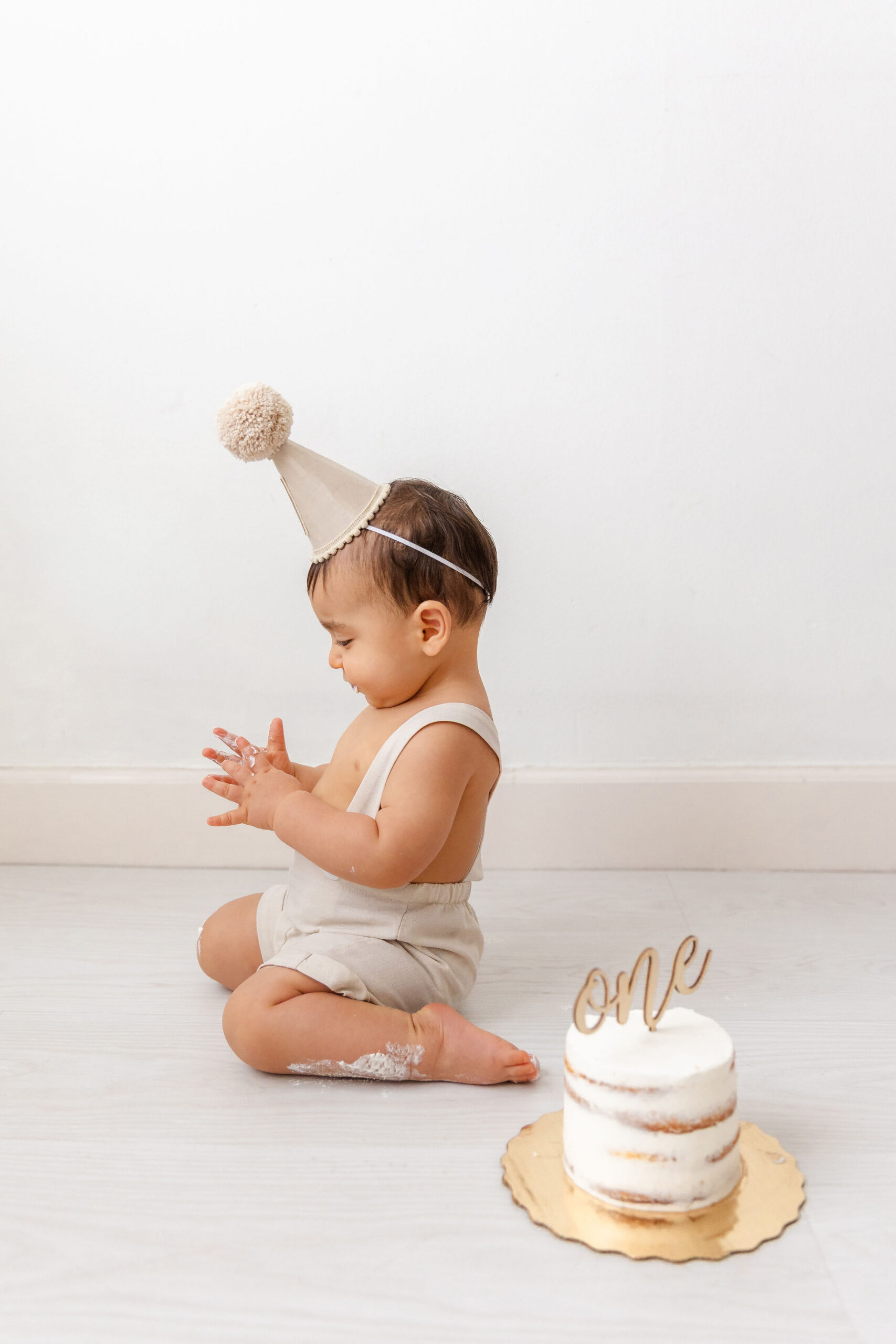 A happy baby claps in tan overalls and birthday cap behind a first birthday cake after loving indoor playgrounds in coral gables