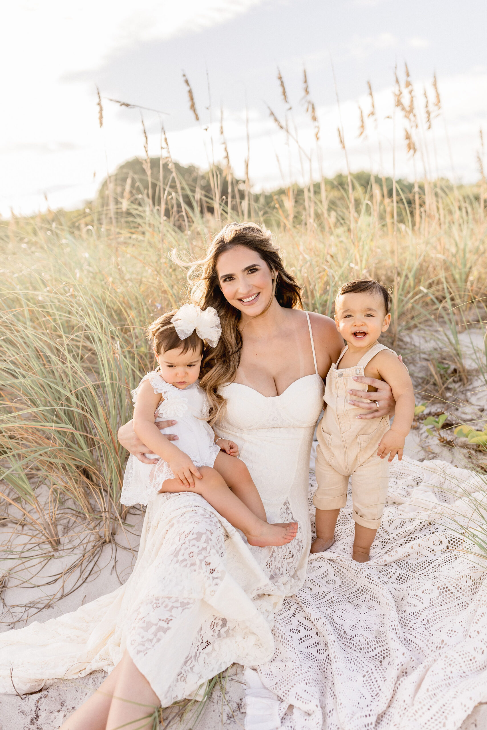 A smiling mother sits on a beach at sunset with a daughter and her twin brother with her