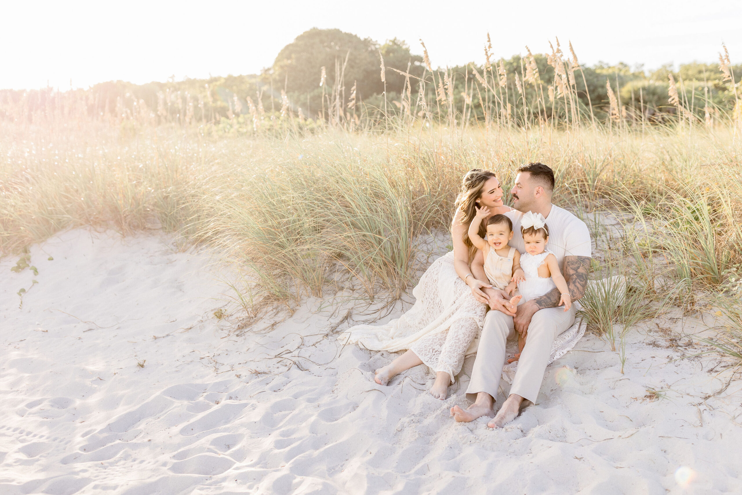 A mom and dad smile at each other while sitting in a beach dune at sunset with happy twin toddlers in their laps after exploring montessori schools in coral gables