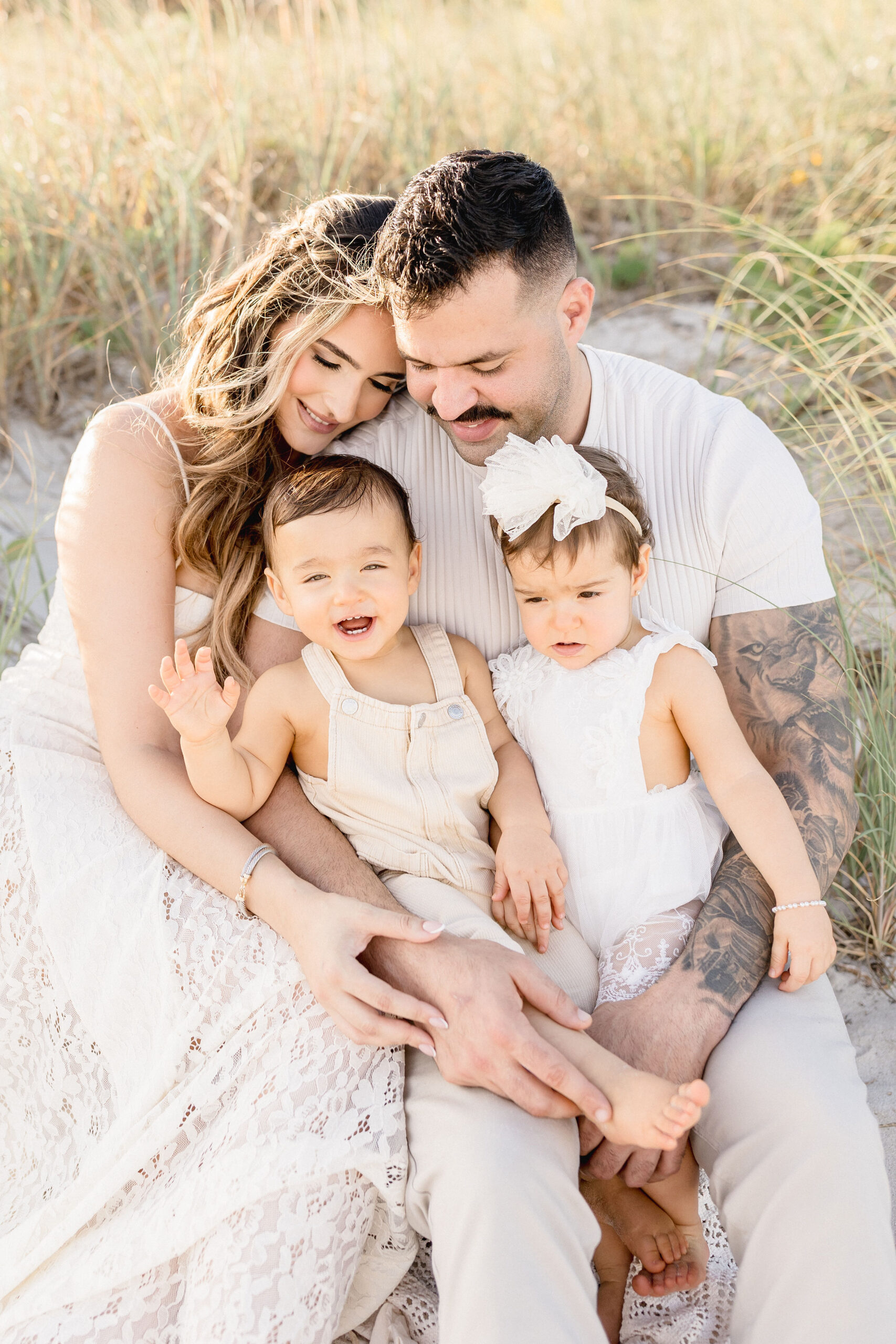 Happy mom and dad sit on the beach in the dunes with their twin toddlers in white in their laps after exploring montessori schools in coral gables
