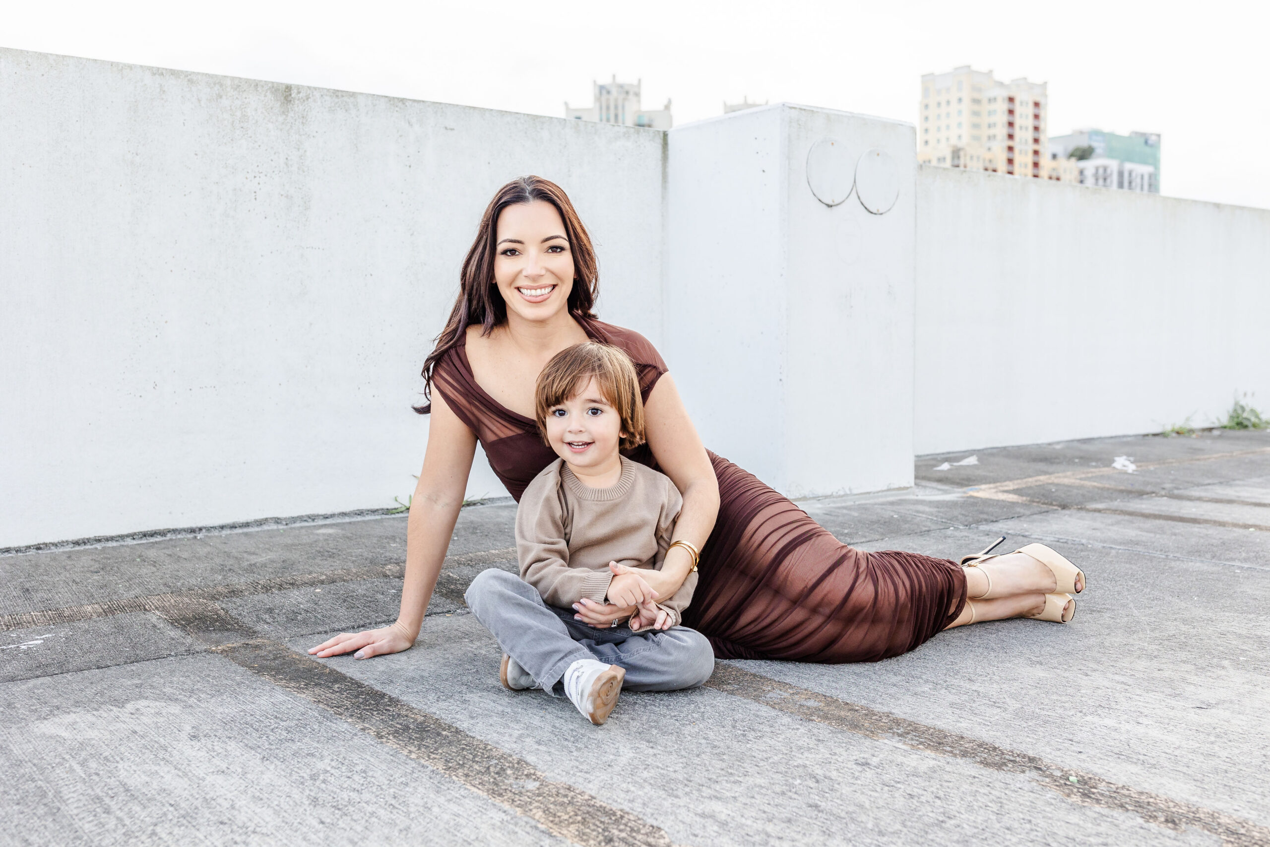 A smiling mother in a brown dress sits across the ground on a parking garage rooftop with her toddler son smiling