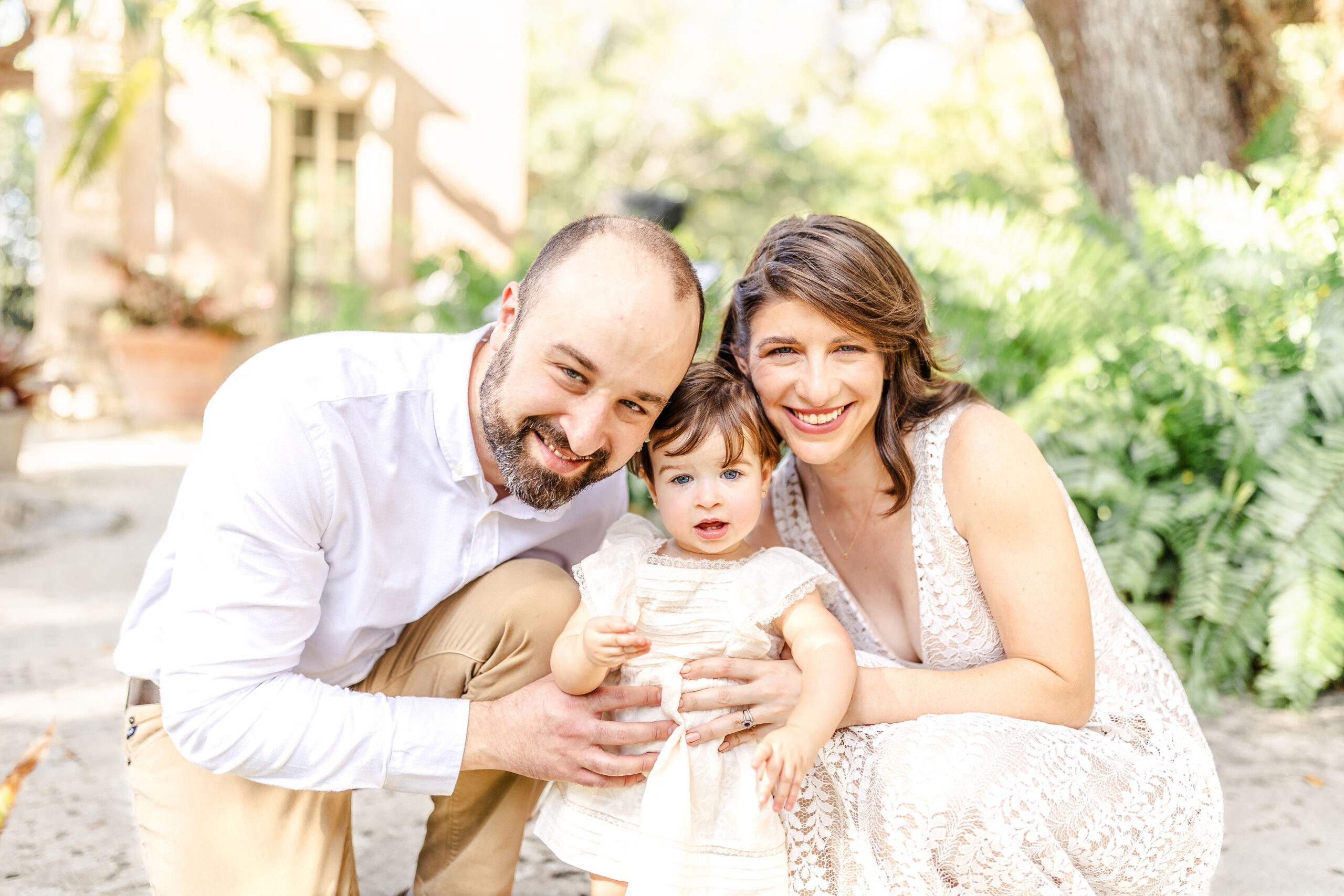 A mother and father kneel to hug their happy toddler daughter in a tropical garden