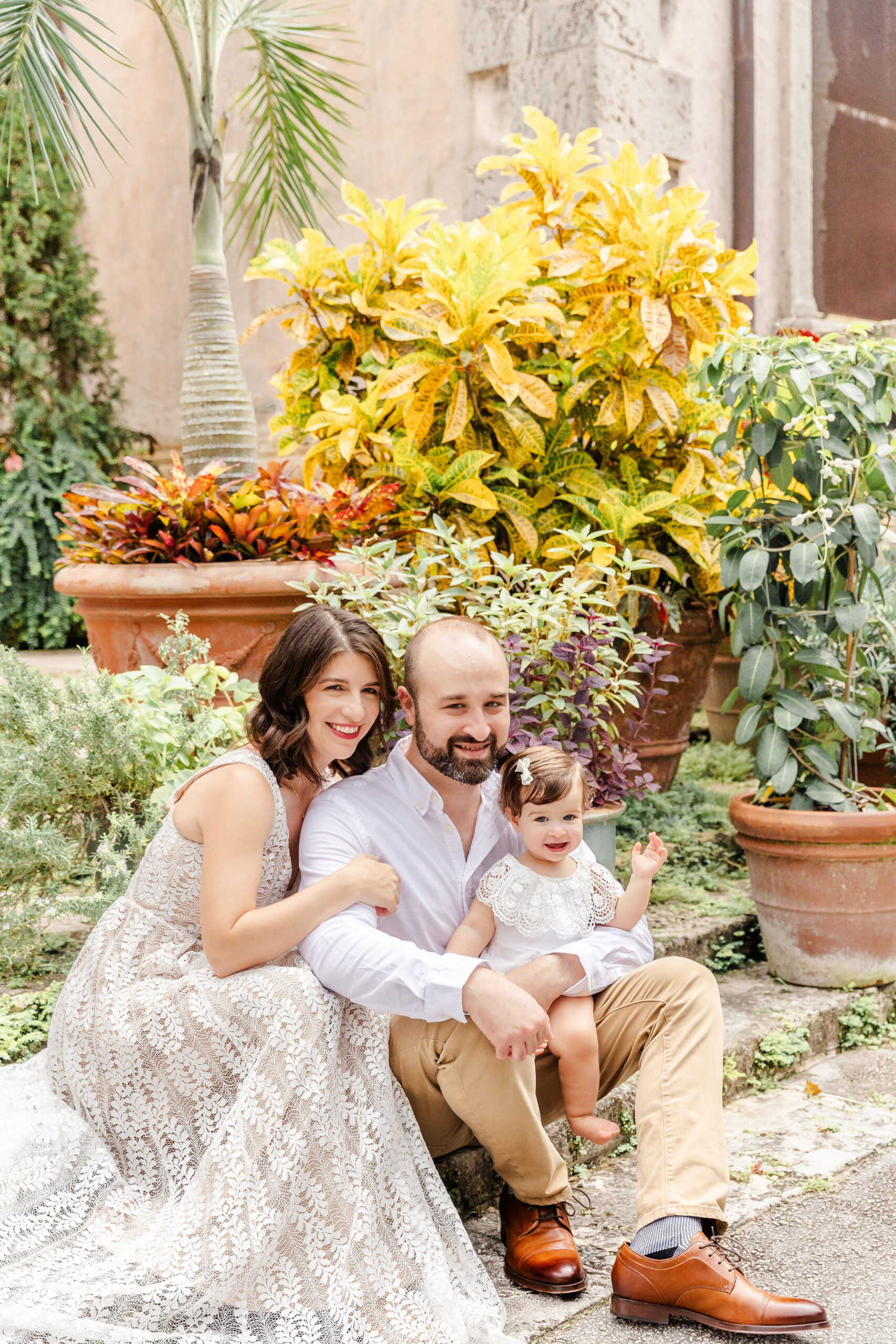 A happy mom and dad sit in a garden with their smiling toddler daughter in their lap after finding great things to do in coral gables with kids