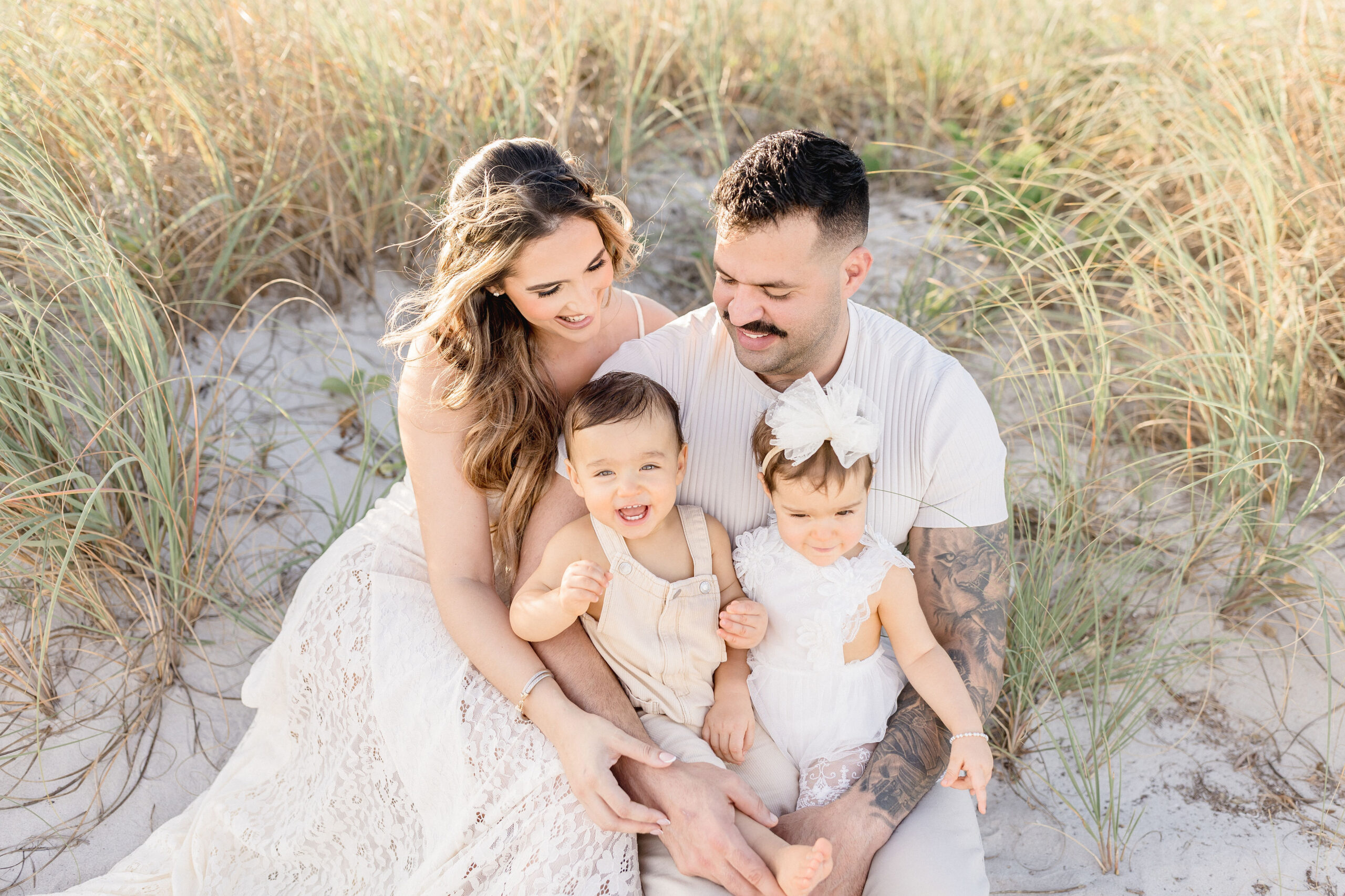 Happy mom and dad sit in a beach dune at sunset with heir giggling toddler twin son and daughter in their laps