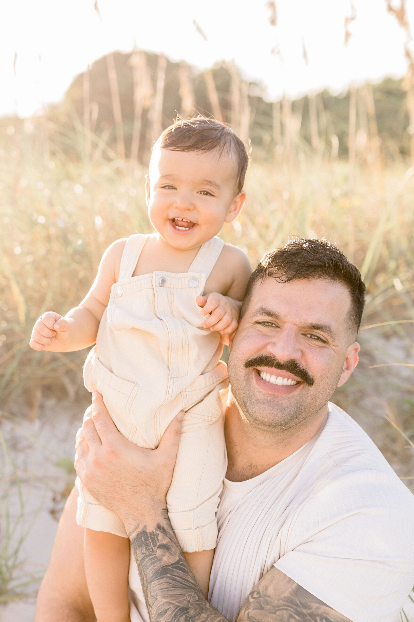 A proud da lifts his giggling toddler son in cream overalls on a beach at sunset after finding spots for kids haircuts in miami