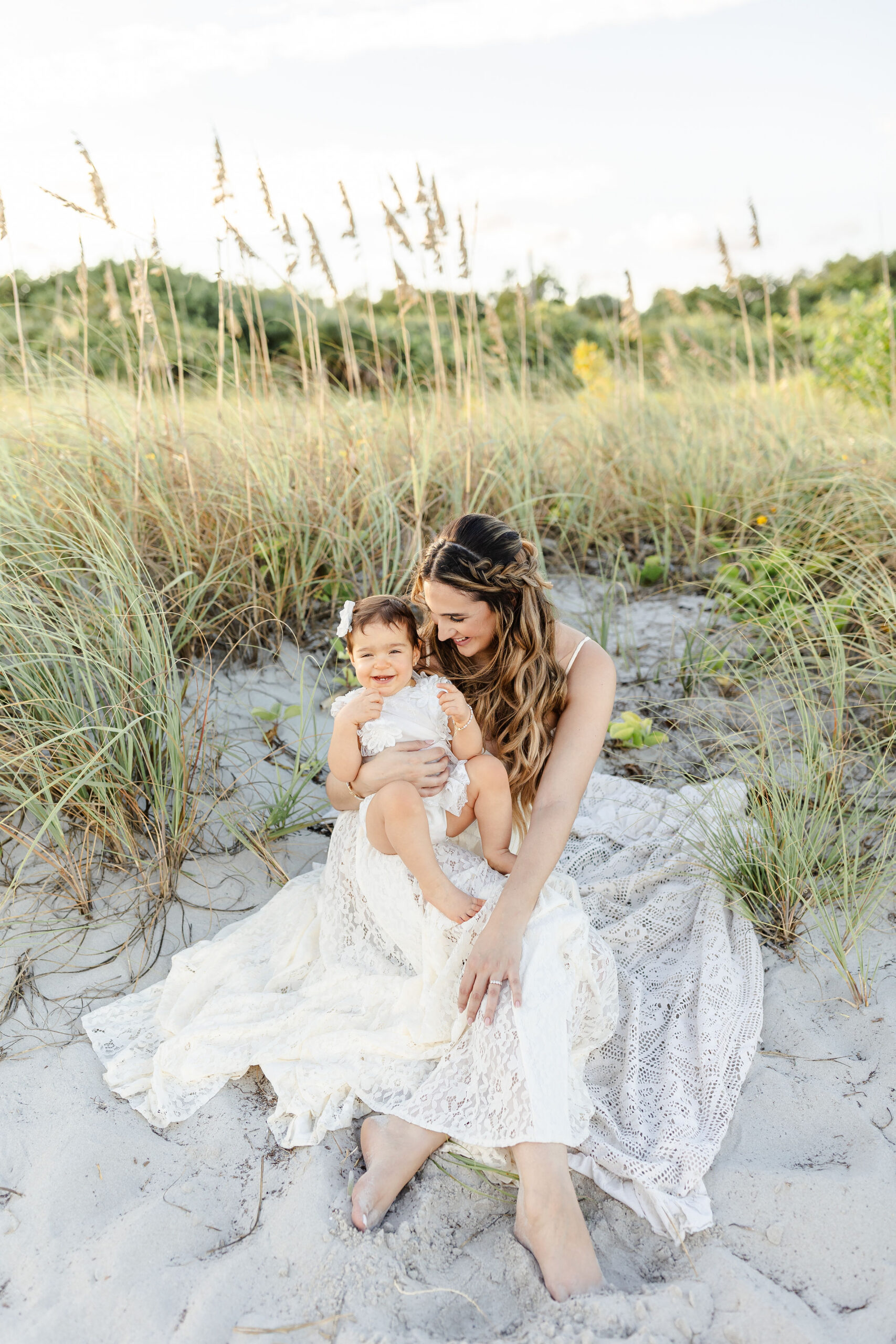 A toddler girl in white giggles in mom's lap on a beach at sunset on a picnic blanket after finding spots for kids haircuts in miami