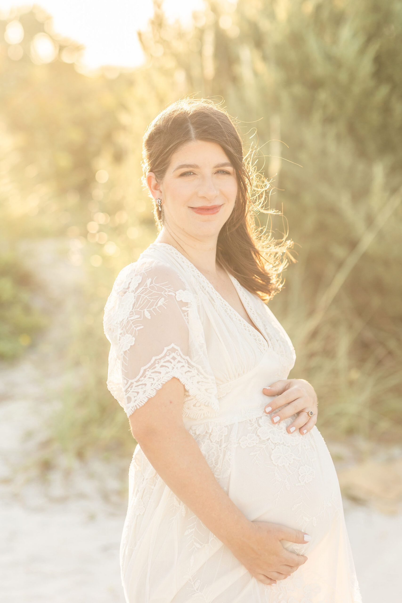 A smiling mother to be in a lace maternity dress stands on a beach at sunset with ahnds on the bump