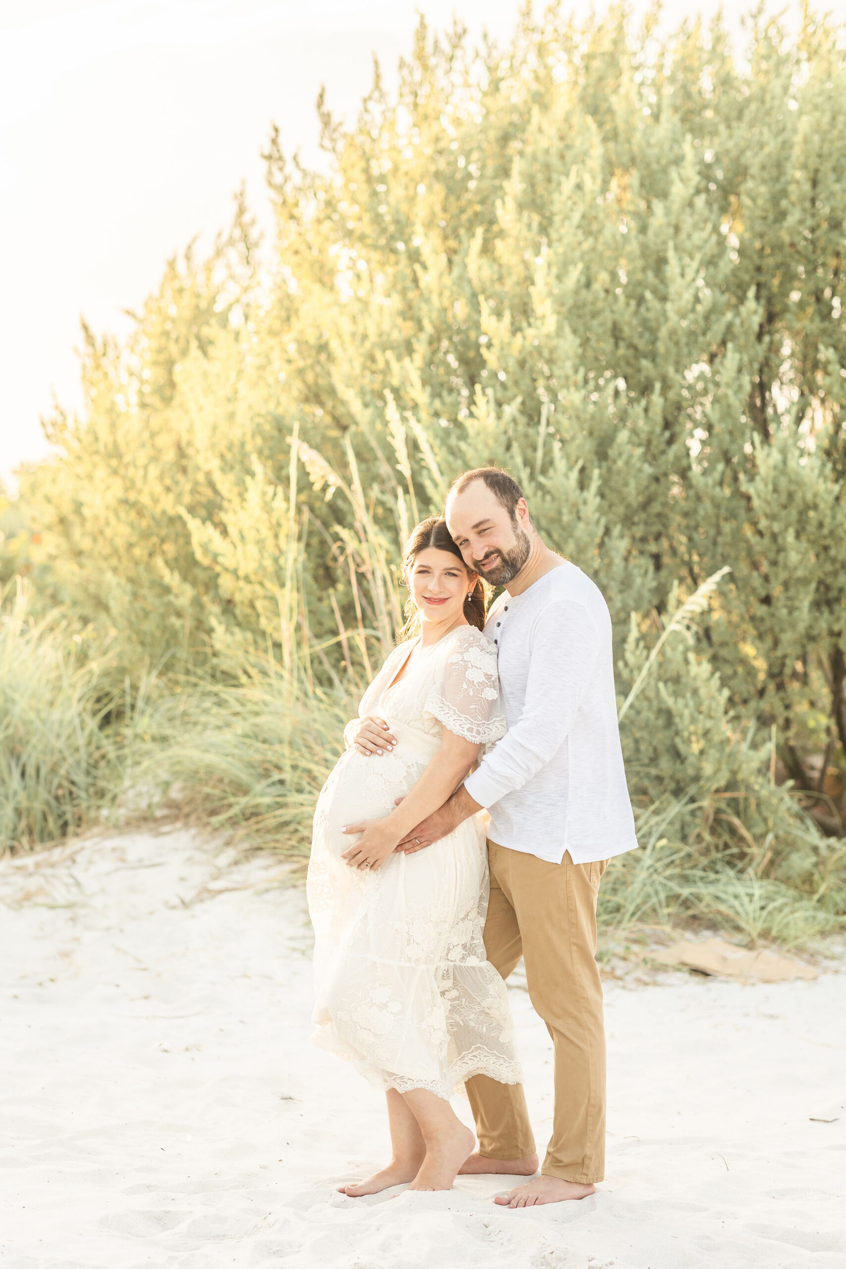 A pregnant couple snuggles and smiles on a beach with hands on the bump in cream and white after finding mat and maternity