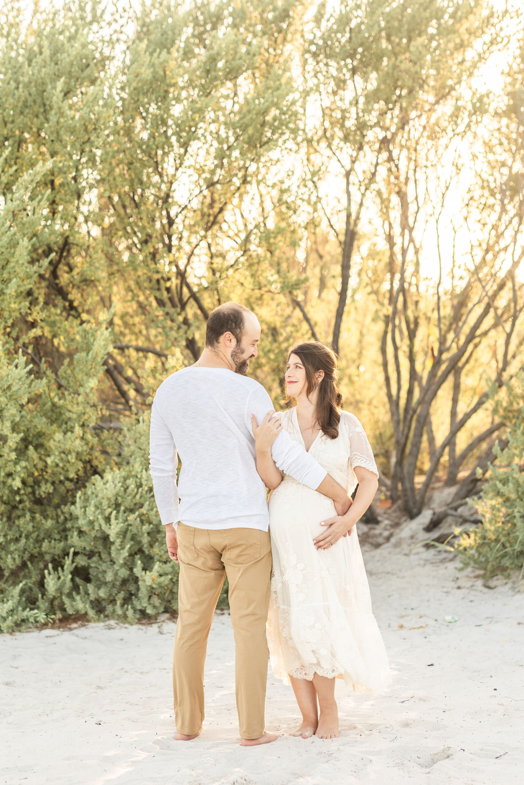 A happy expecting couple stand on a beach in white at sunset smiling at each other with arms wrapped after visiting mat and maternity