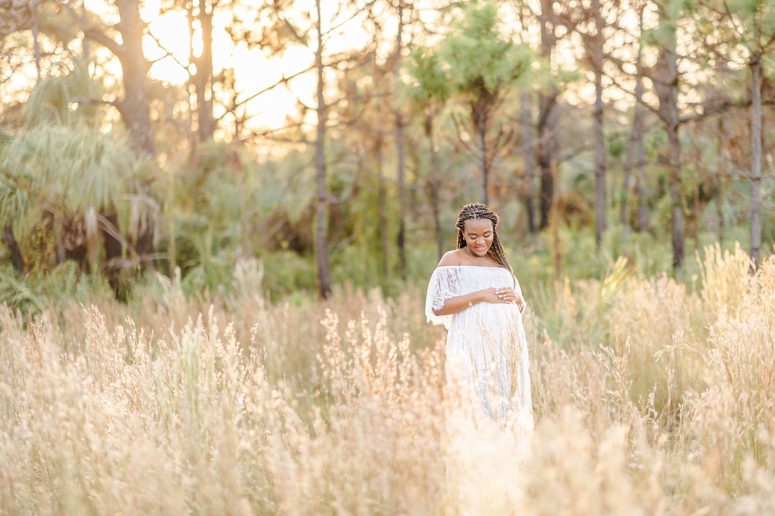An expectant woman in a white lace maternity gown stands in tall golden grass at sunset