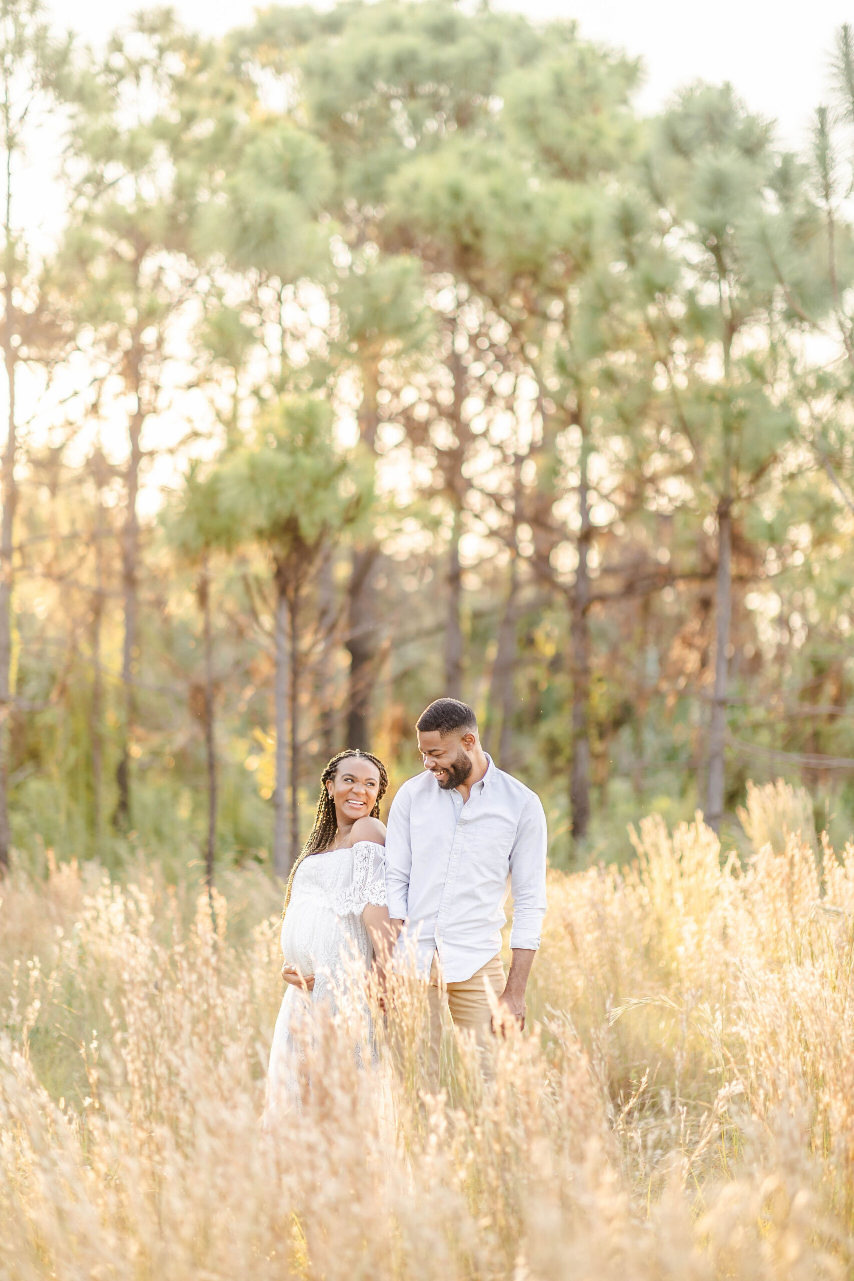 A smiling mother to be in a white lace dress walks in tall golden grass at sunset with her husband holding hands at sunset after finding miami mami