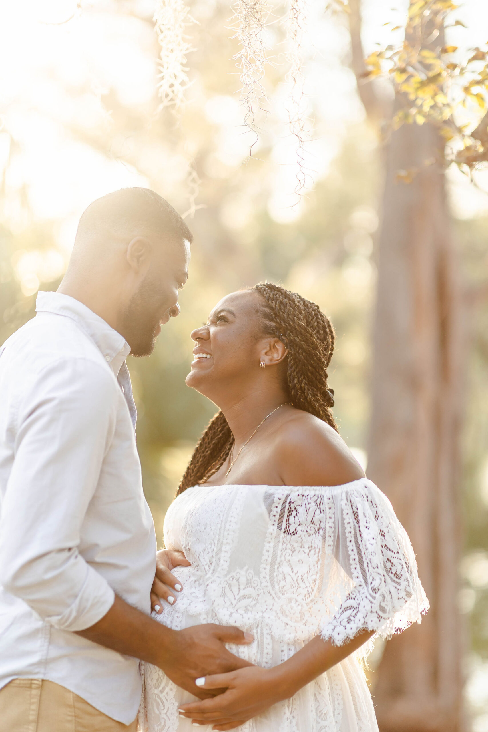 A happy pregnant couple laughs while holding the bump in a lace white maternity gown in a park after visiting miami mami
