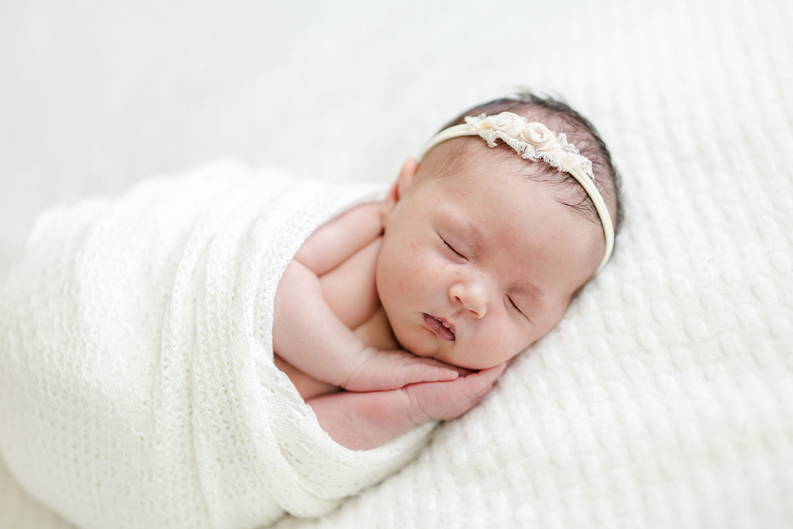 A newborn baby girl in a flower headband sleeps on her side on her hands in a white knit swaddle after visiting Minnie Miami Beach