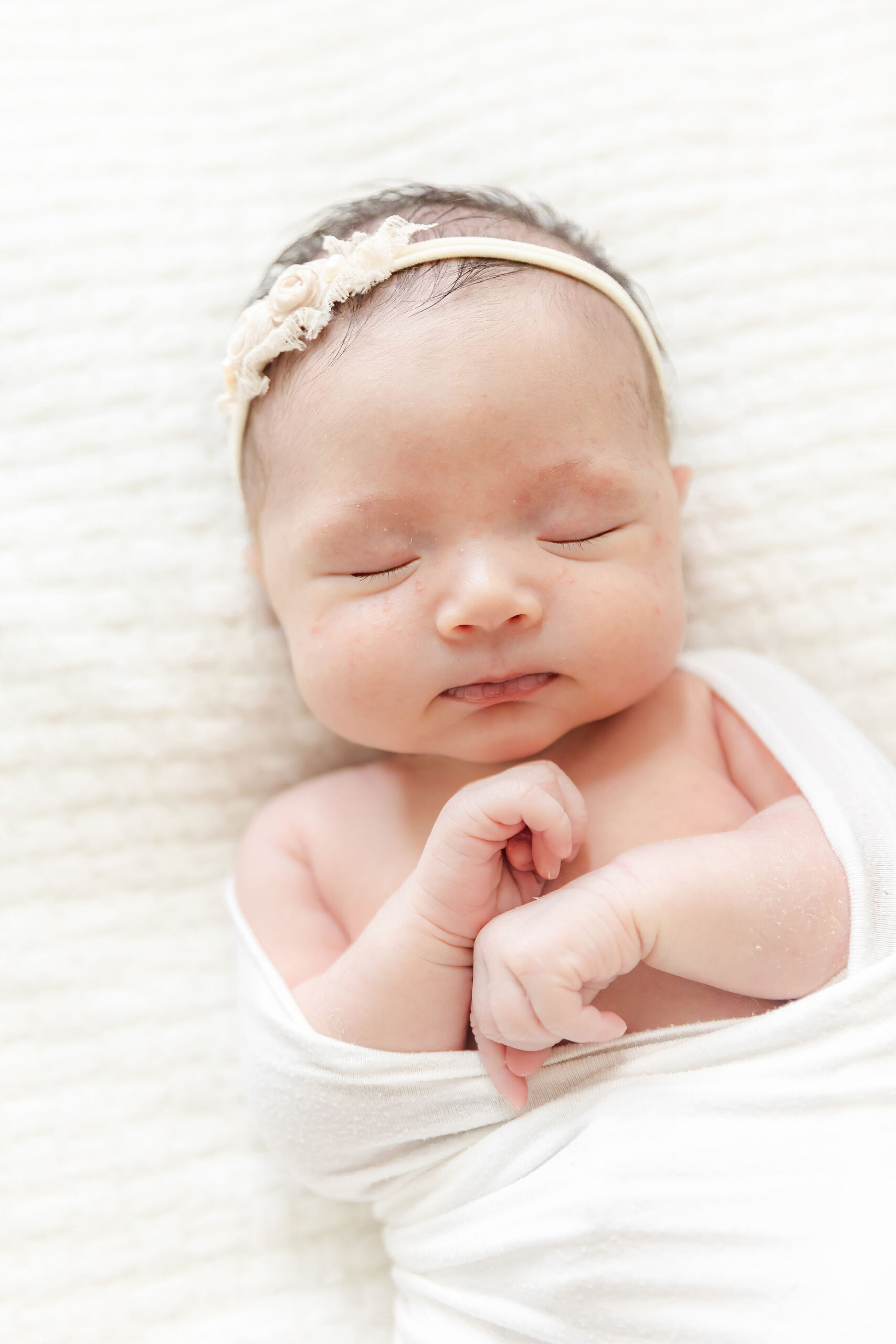 A newborn baby girl sleeps in a white slightly open swaddle and headband on a white bed after visiting Minnie Miami Beach