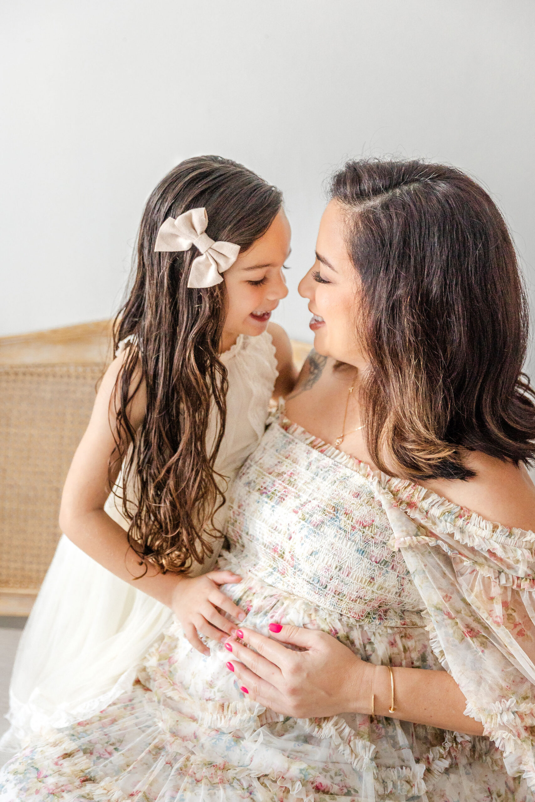 A happy toddler girl in white leans in to kiss mommy while sitting on a bench in a studio in floral dresses and hands on the bump