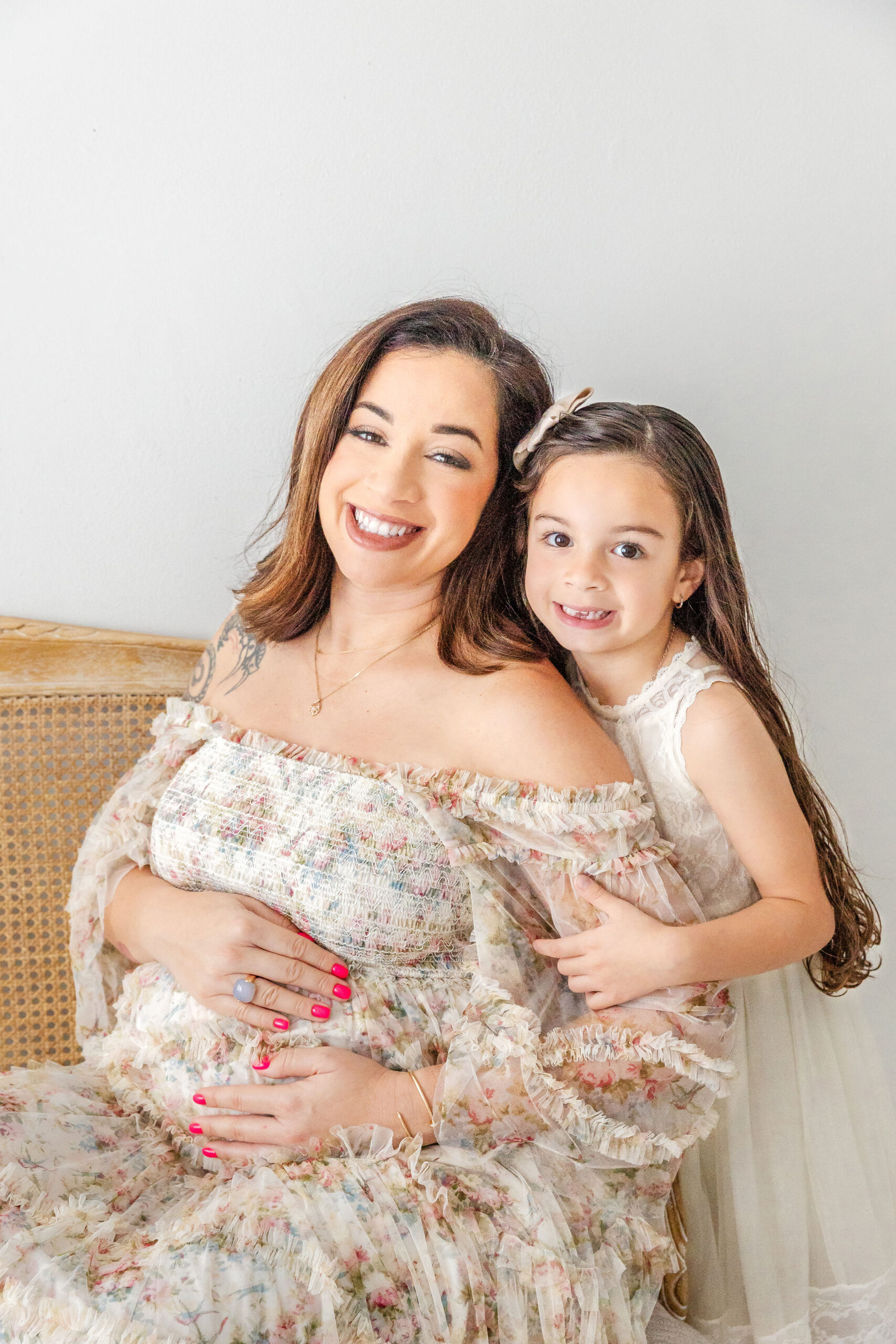 A happy expectant mother and her toddler daughter smile big while in floral print and white lace dresses in a studio after meeting Werk It Moms