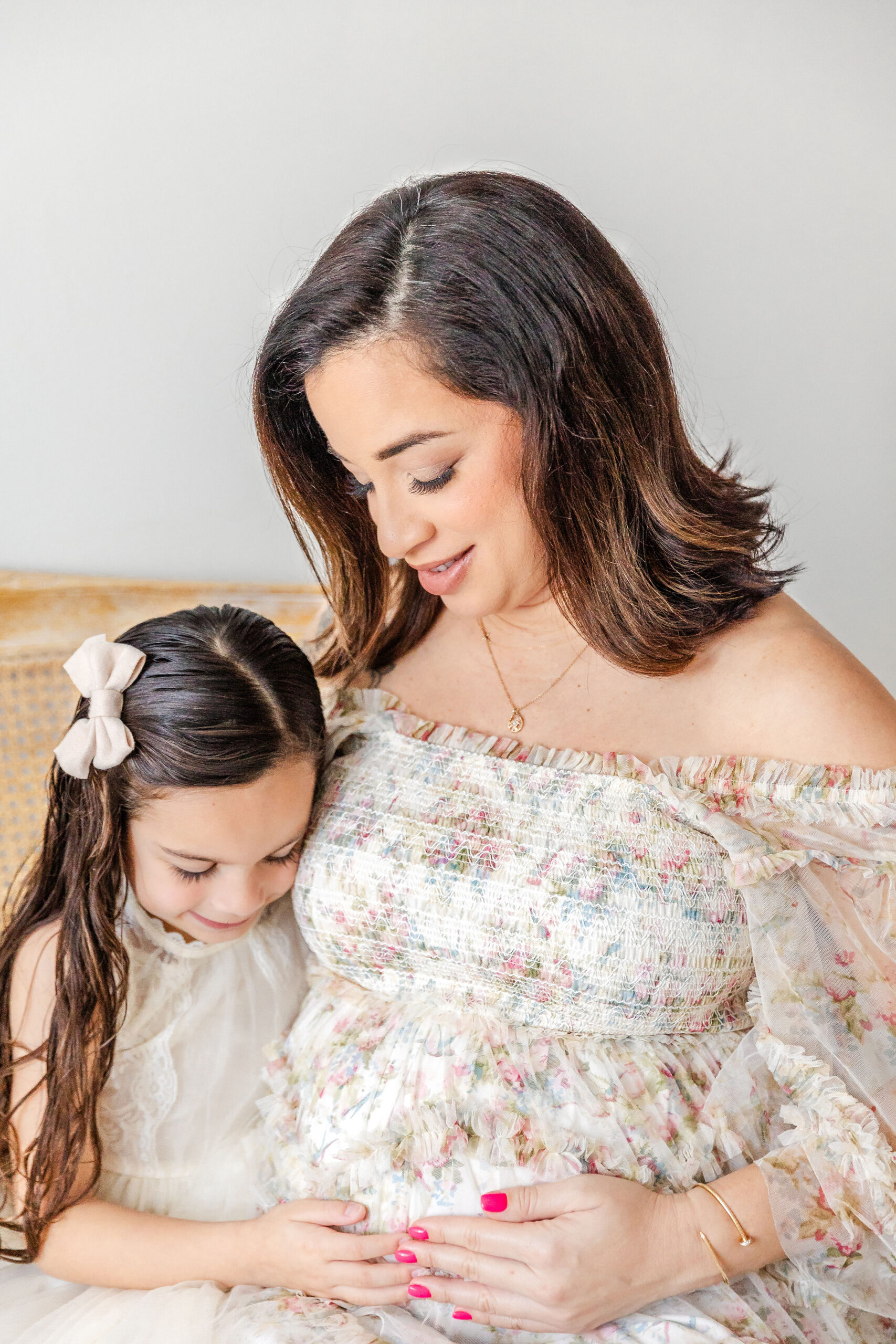 A happy pregnant mother in a floral print dress sits holding her bump with her toddler daughter in white smiling down to it after visiting Werk It Moms