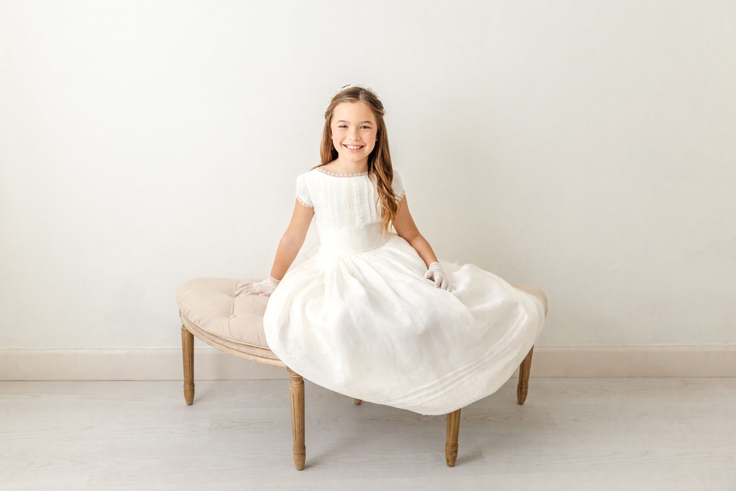 A happy young girl sits across a bench with white gloves and a white first communion dress found at cyberella baby