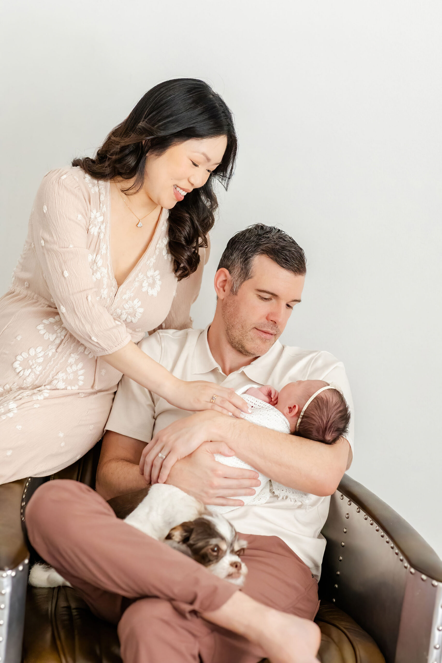 A new dad sits in a chair cradling his newborn daughter in his arms as mom reaches in sitting on the armrest
