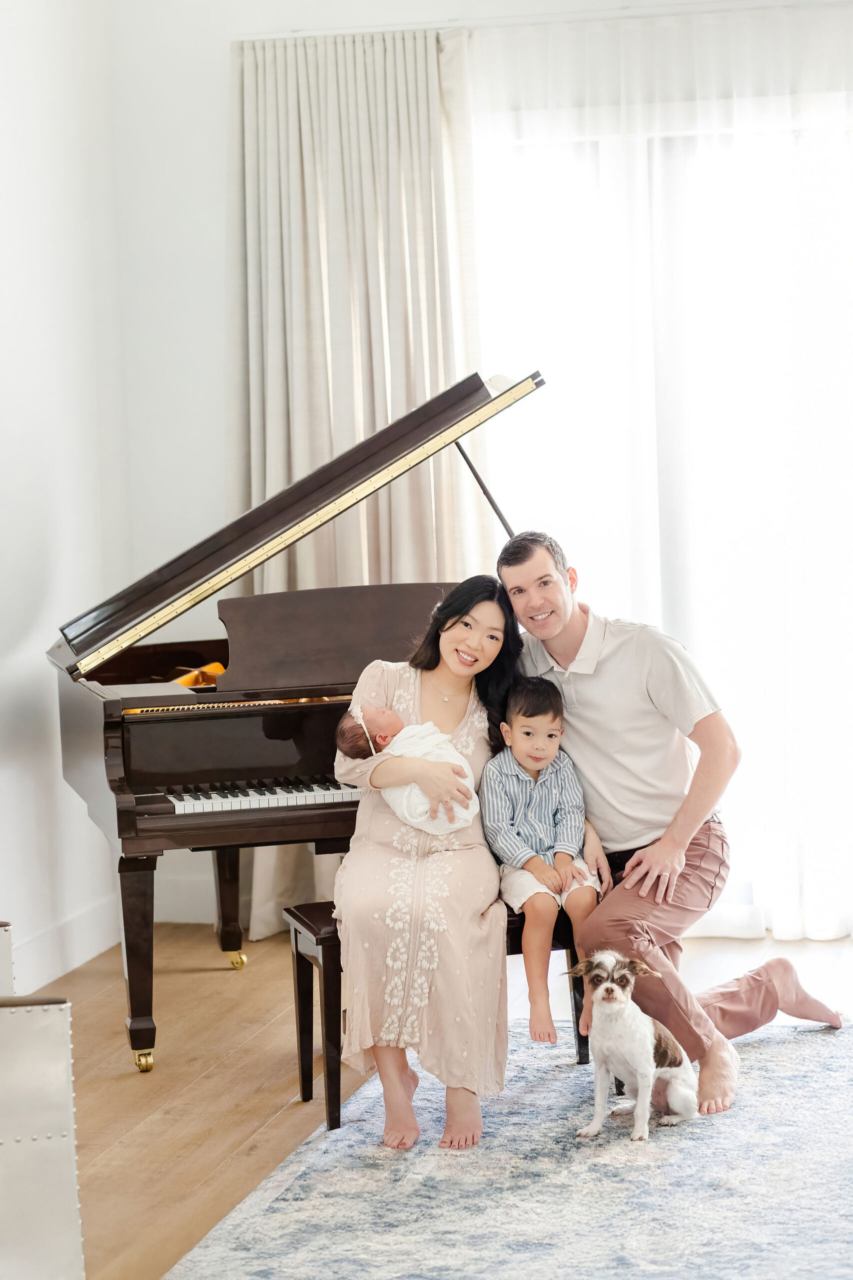 A happy mom and dad sit on a piano bench holding their newborn and toddler son in the middle after meeting harmony baby concierge