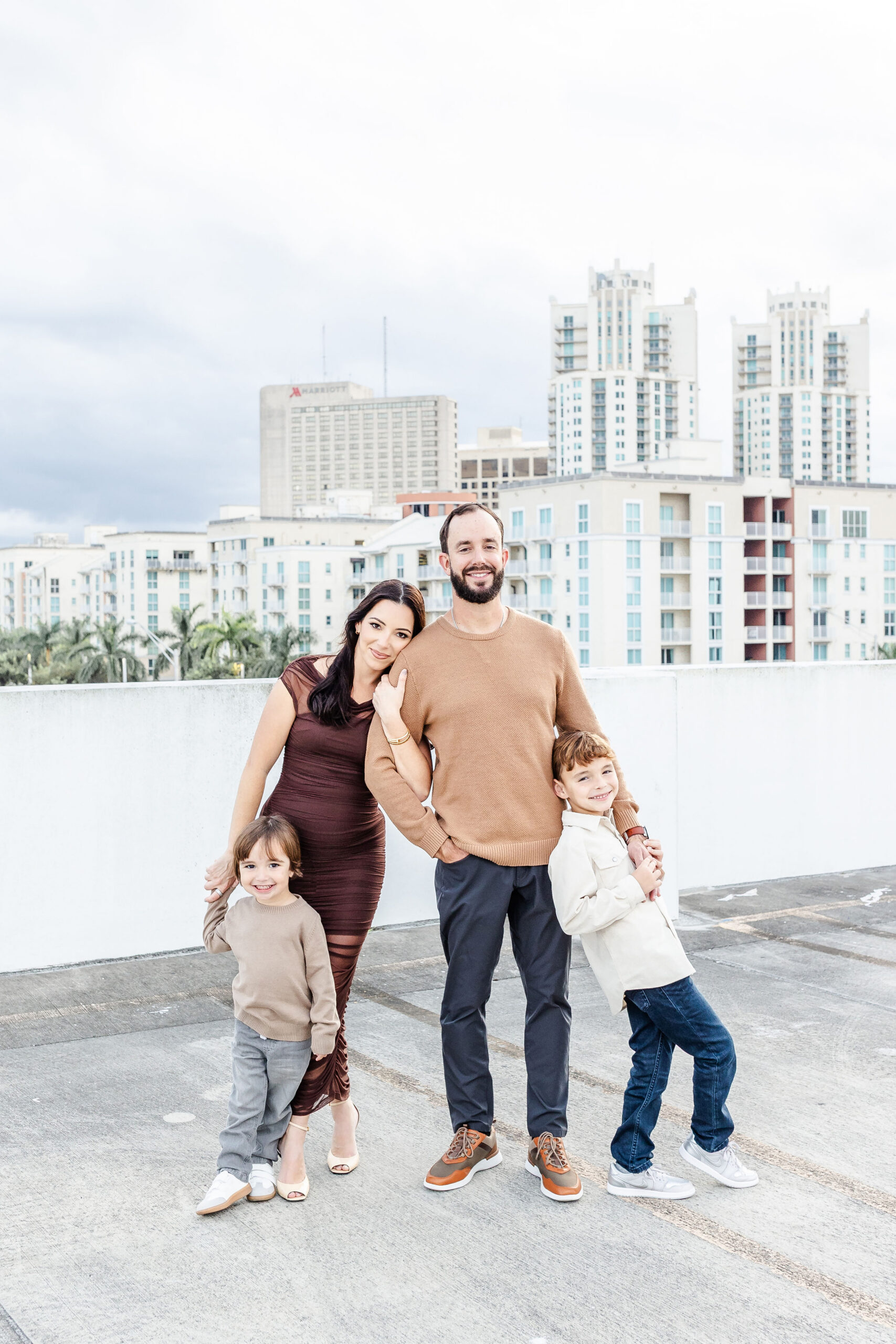 Happy mom leans on her husband as they stand with their two toddlers in downtown miami on a parking garage roof
