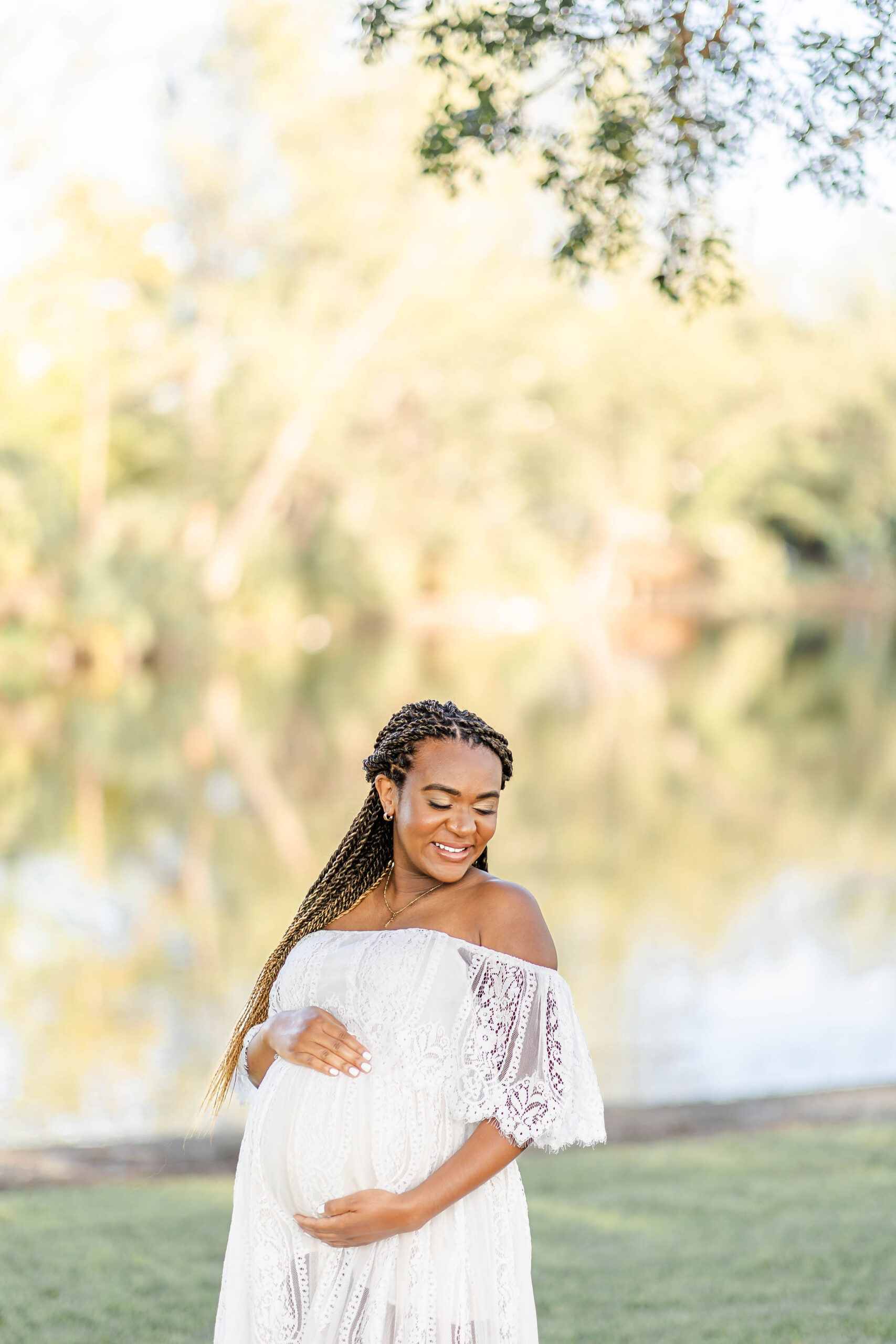 A mother to be smiles down her shoulder while standing in a lace gown by a lake after meeting with mommy n' me doula