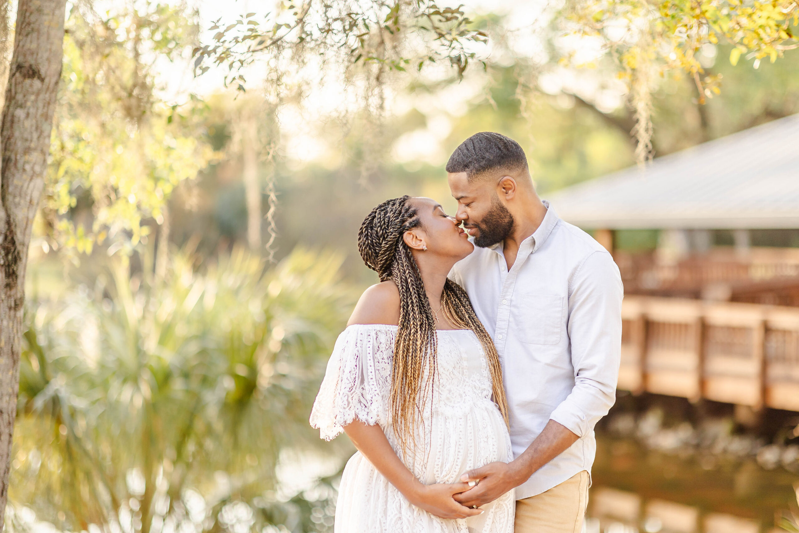 Happy parents to be in white stand by a lake at sunset leaning in for a kiss with hands under bump after meeting mommy n' me doula