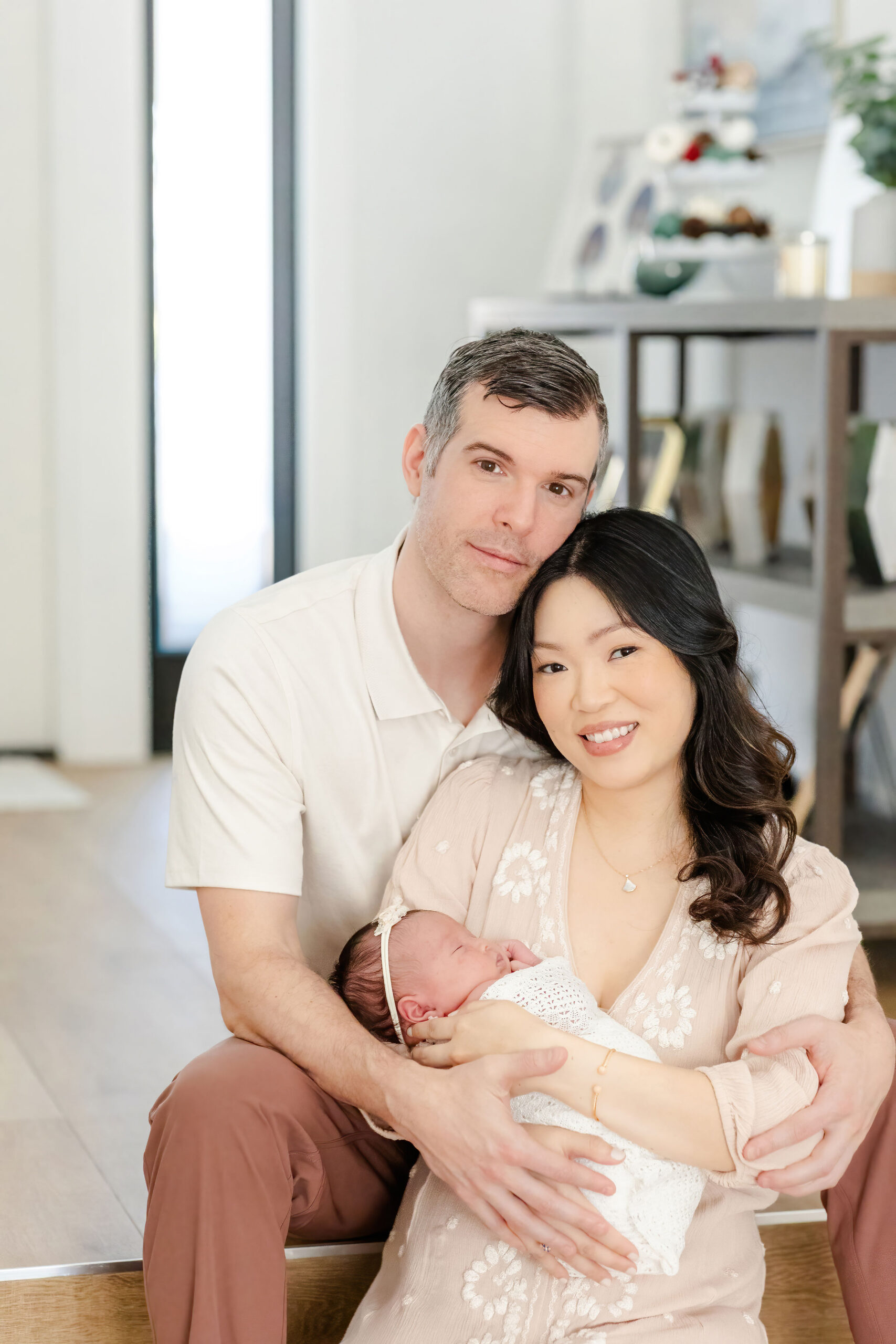 A mom and dad snuggle on steps in their home while holding their sleeping newborn in cream outfits