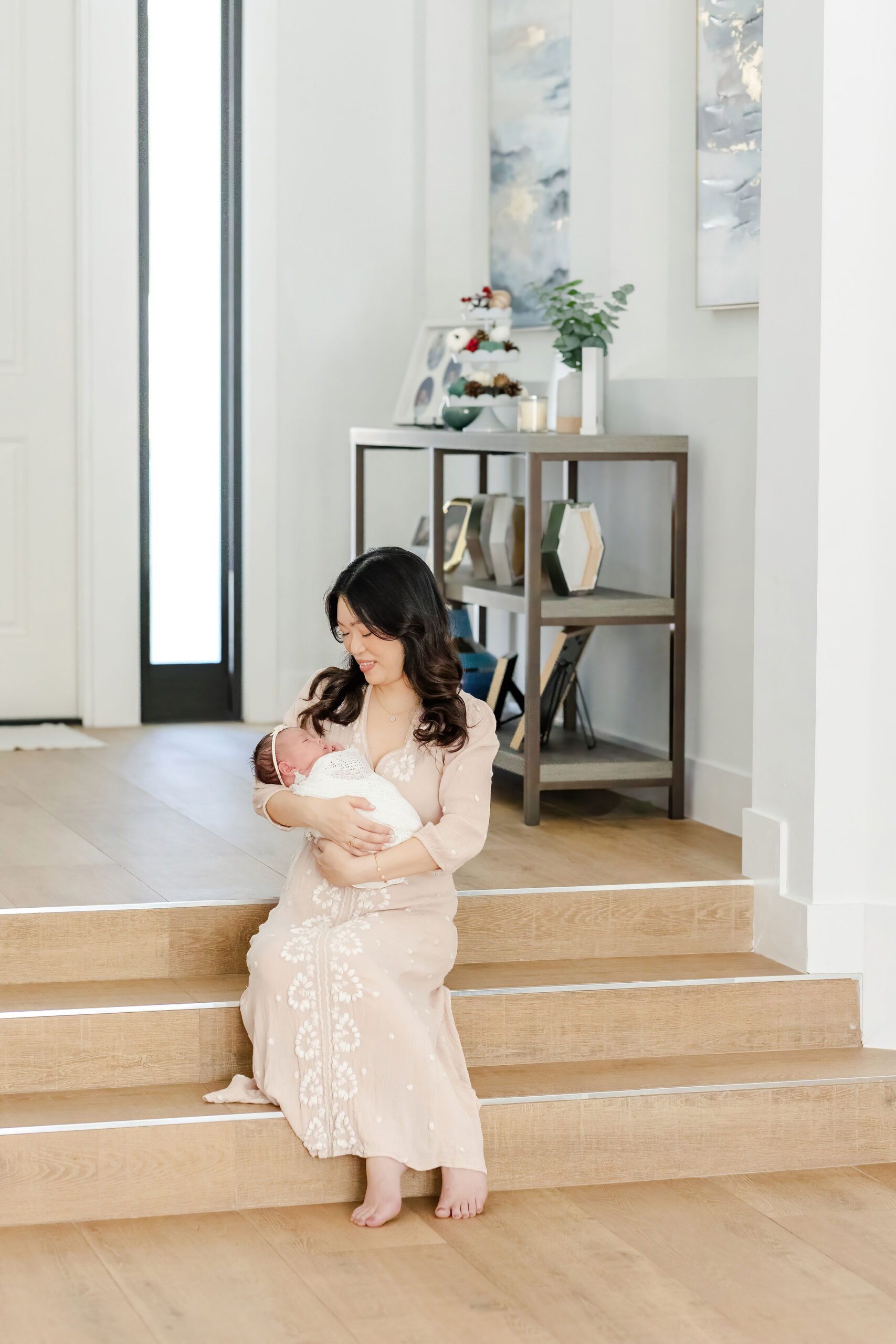 A happy mom sits on wooden house steps cradling her sleeping newborn in her arms in a white swaddle after meeting with my mom glow