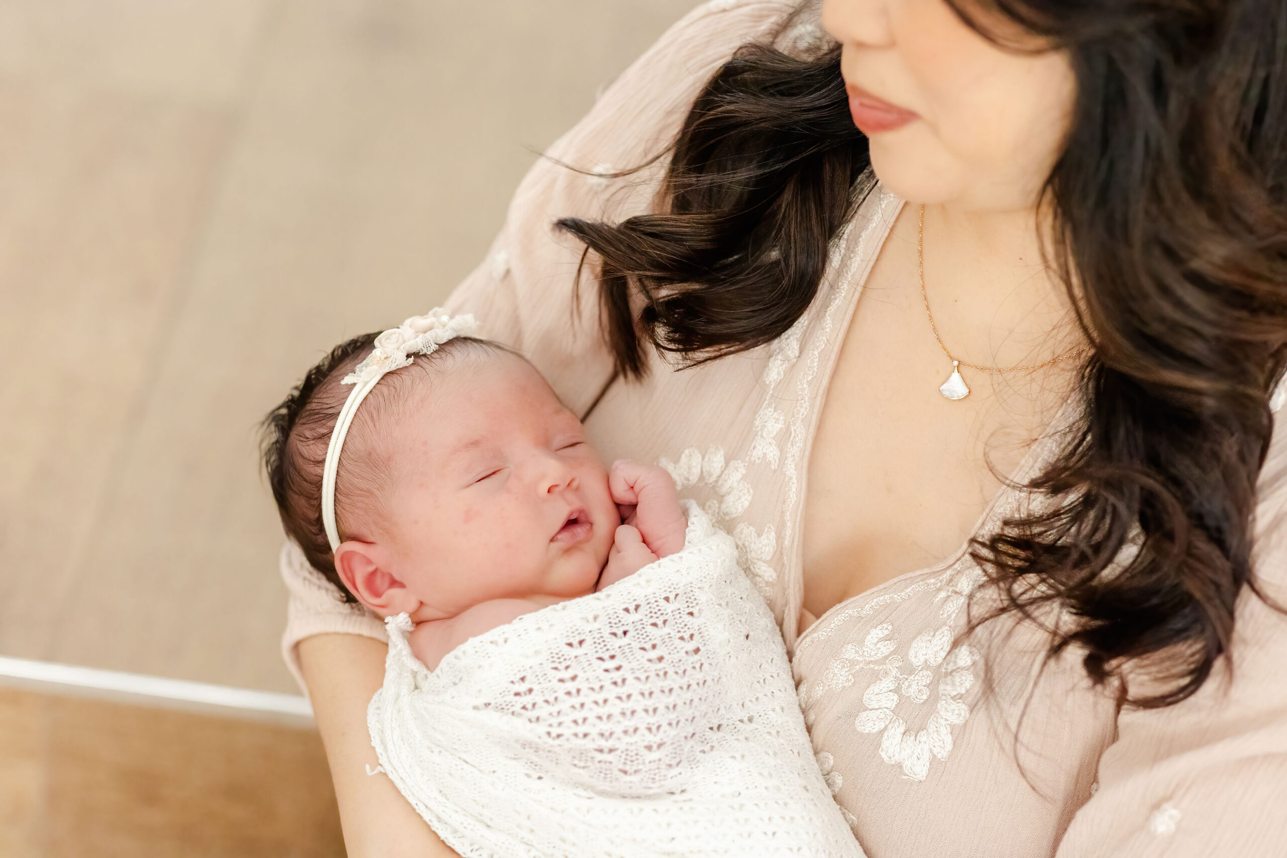 A newborn baby girl in a white knit swaddle sleeps against mom's chest in a studio after visiting my mom glow