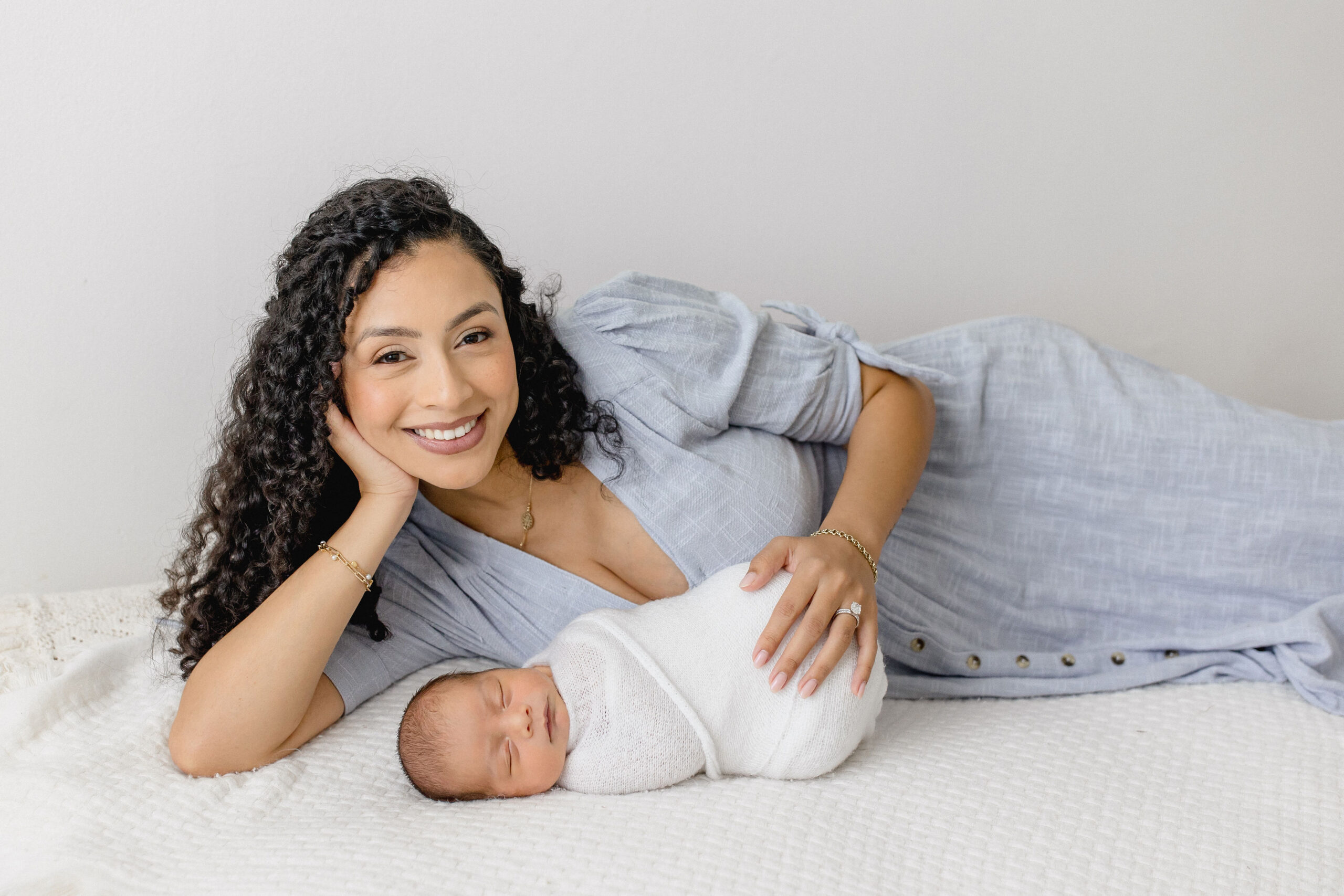 A happy mom in a blue dress lays across a bed with her sleeping newborn in a white swaddle