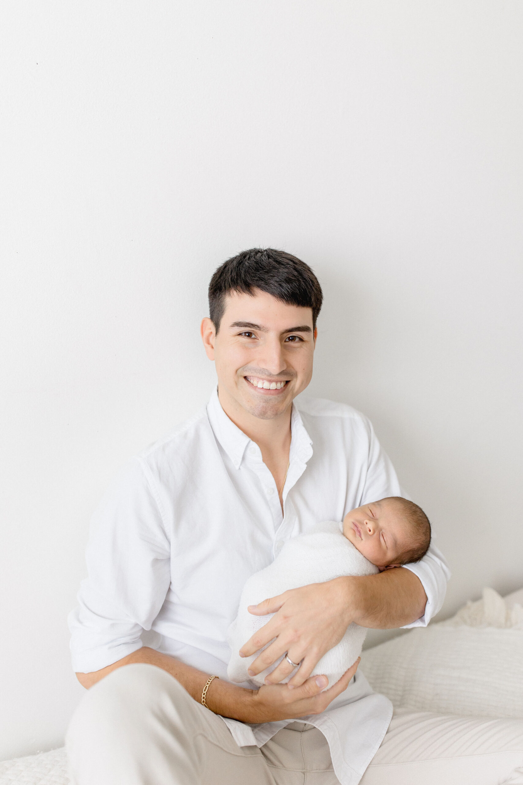 A happy new dad in a white shirt sits on a bed cradling his sleeping newborn in his arms after meeting with twinkle toes nanny agency