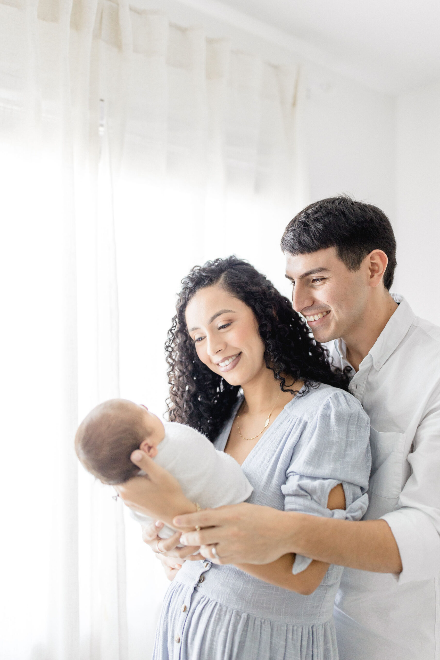 Happy mom and dad stand in a nursery smiling to their newborn in mom's hands after visiting twinkle toes nanny agency