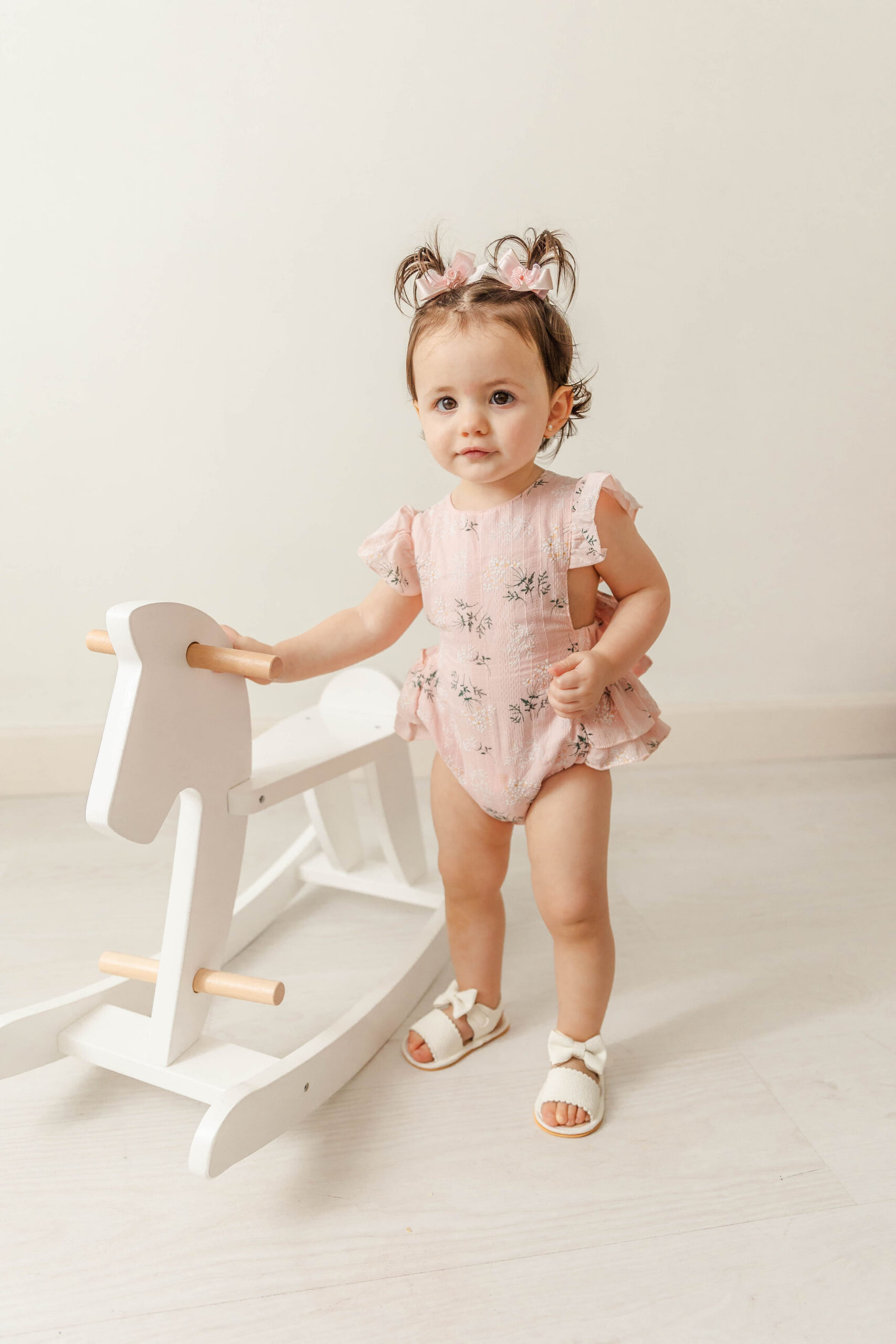 A young toddler girl stands with a wooden sawhorse in a studio in a pink onesie and pigtails after exploring Lilileyba