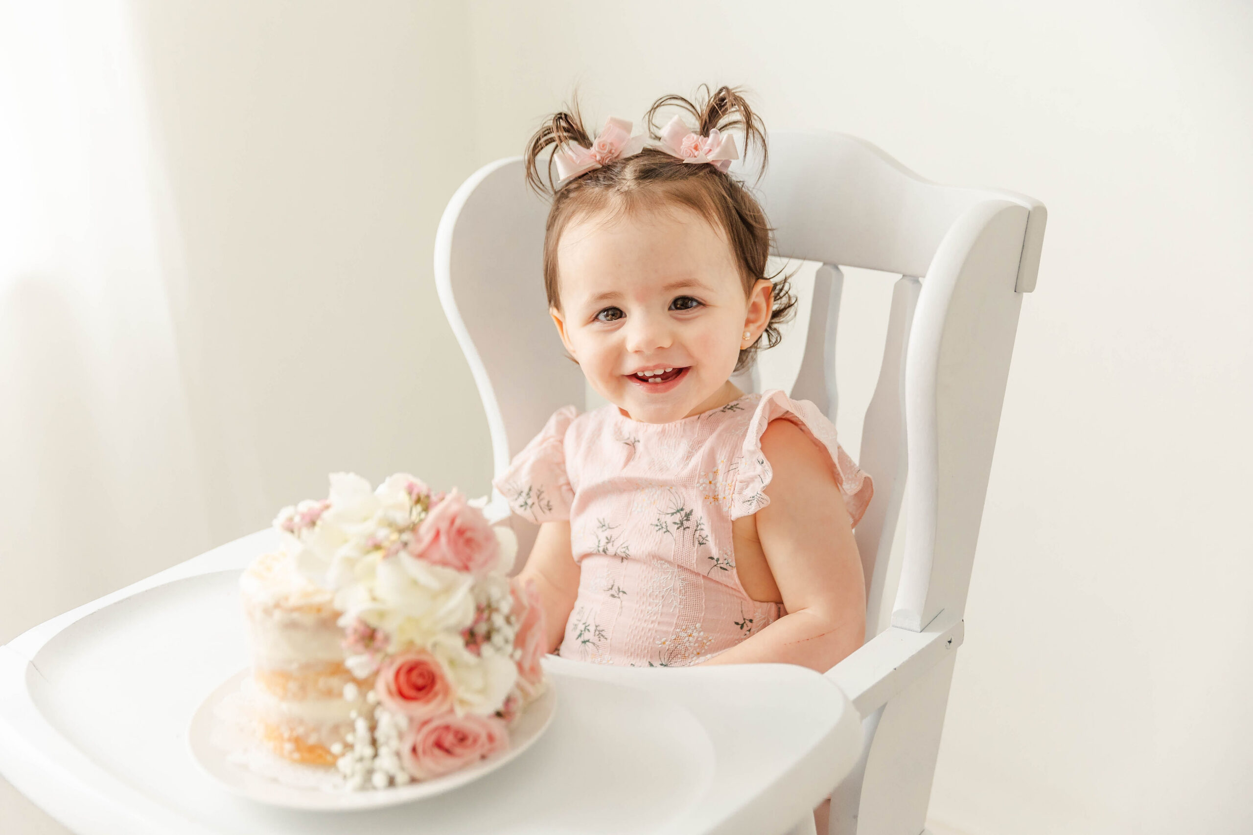 A happy toddler girl in a pink dress smiles while sitting in a high chair behind a flower covered cake after visiting Lilileyba