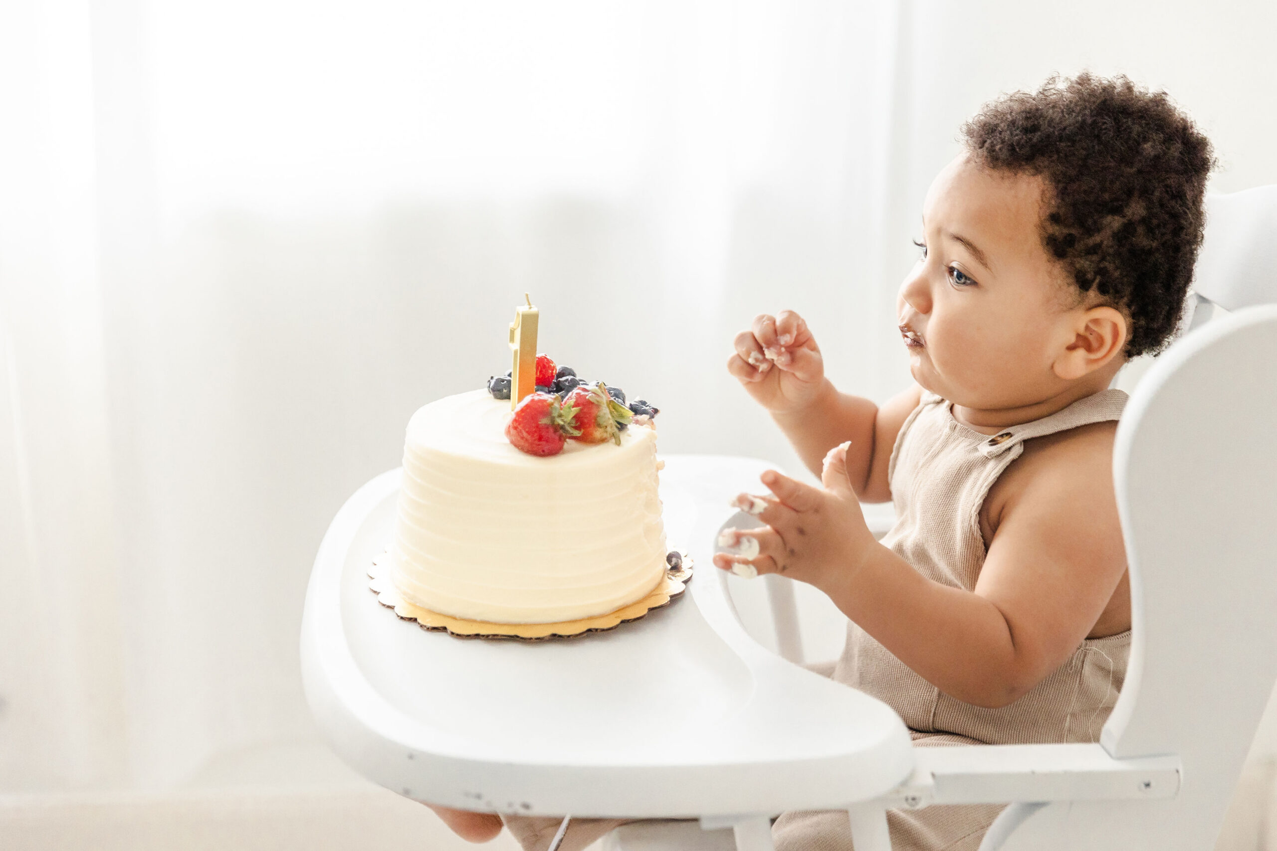 A young toddler boy in brown suspenders reaches for a white fruit covered 1st birthday cake while in a high chair in a studio after finding Pekiboo