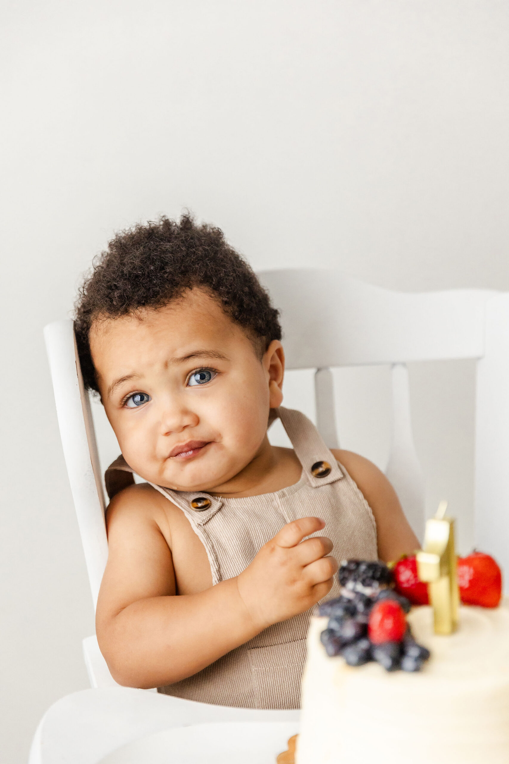 A baby boy in tan suspenders leans on his high chair behind a cake after exploring Pekiboo