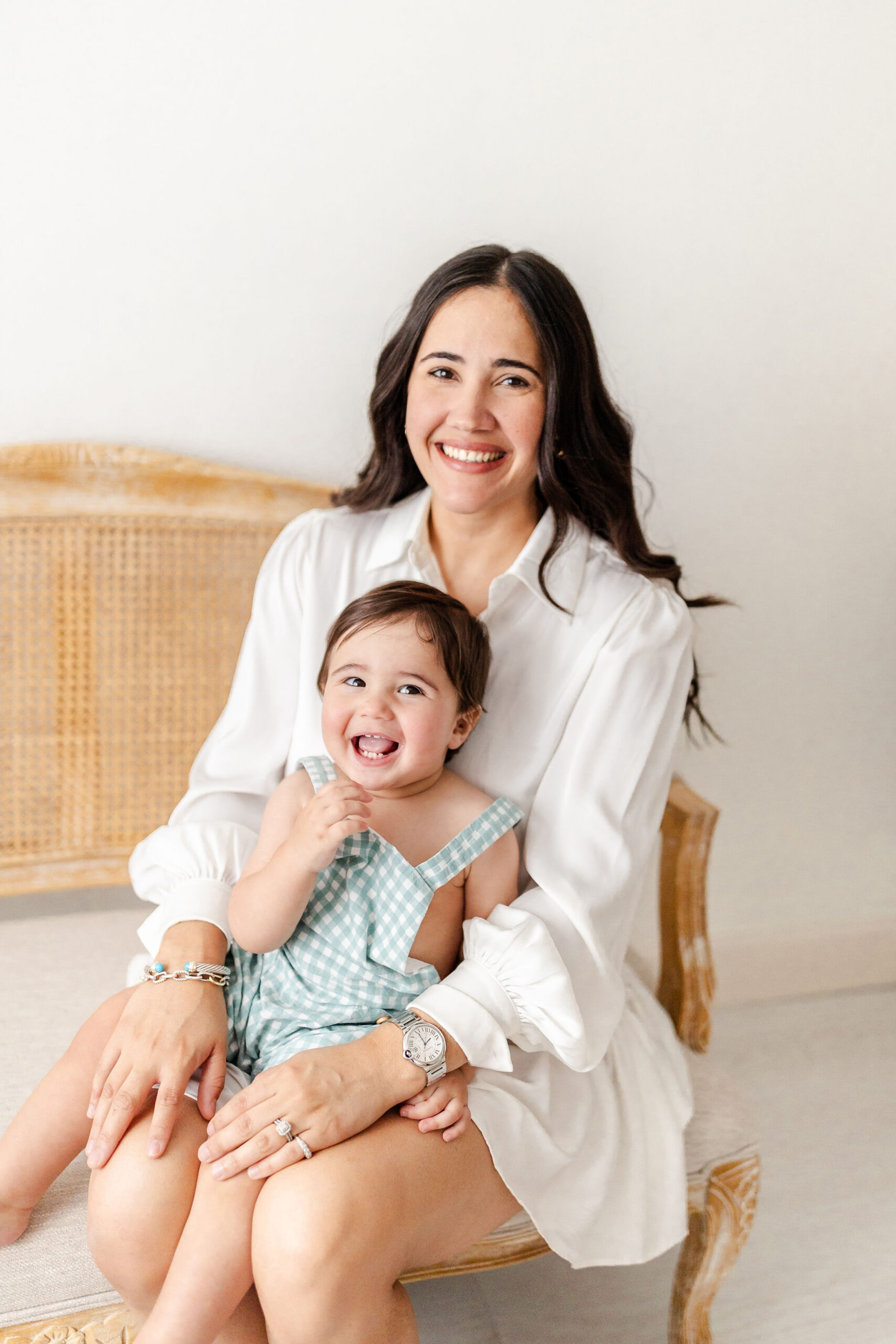 A happy mom in a white dress sits with giggling toddler in her lap on a bench