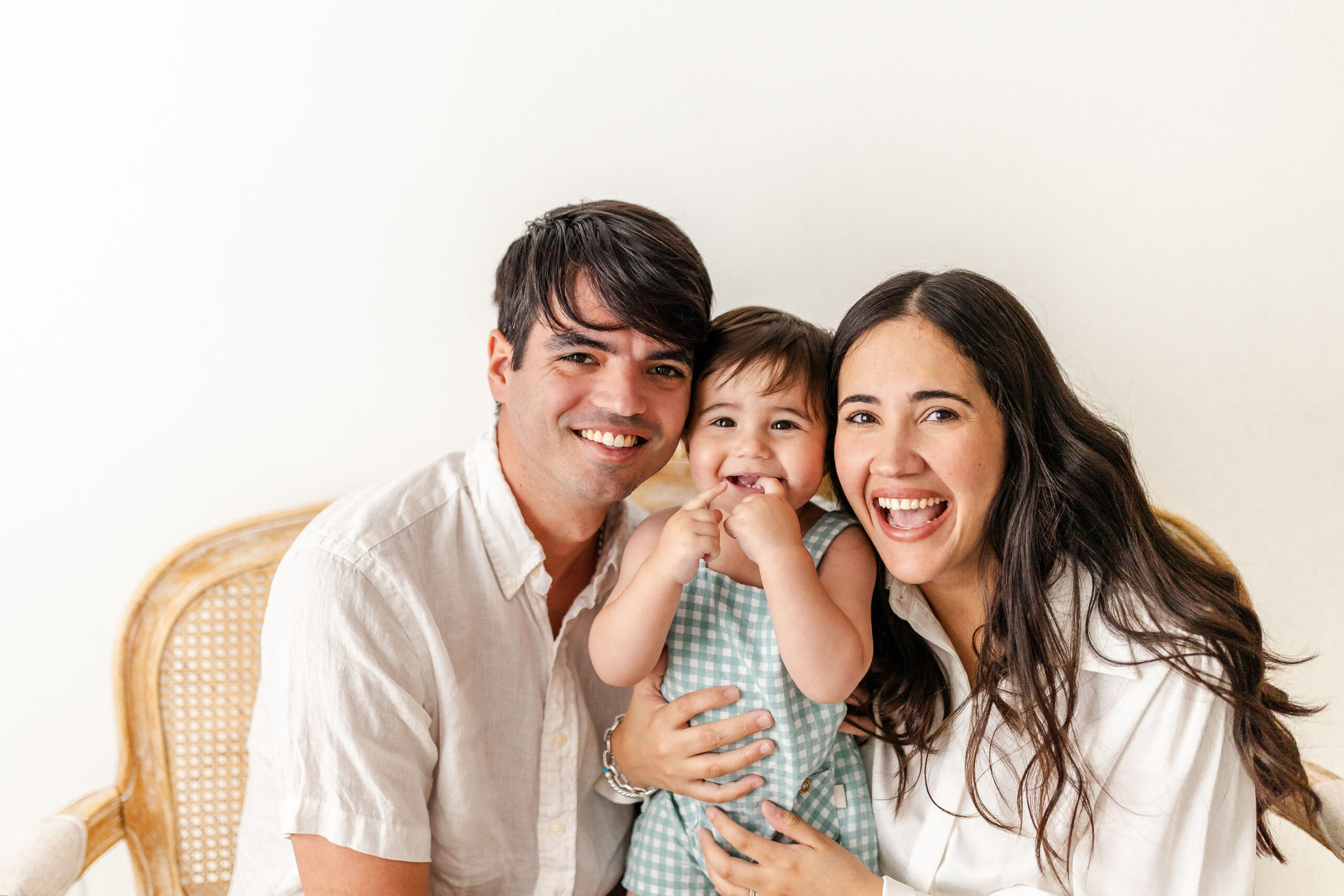 A happy baby in suspenders giggles with fingers in mouth between mom and dad on a bench in a studio after visiting state of kid