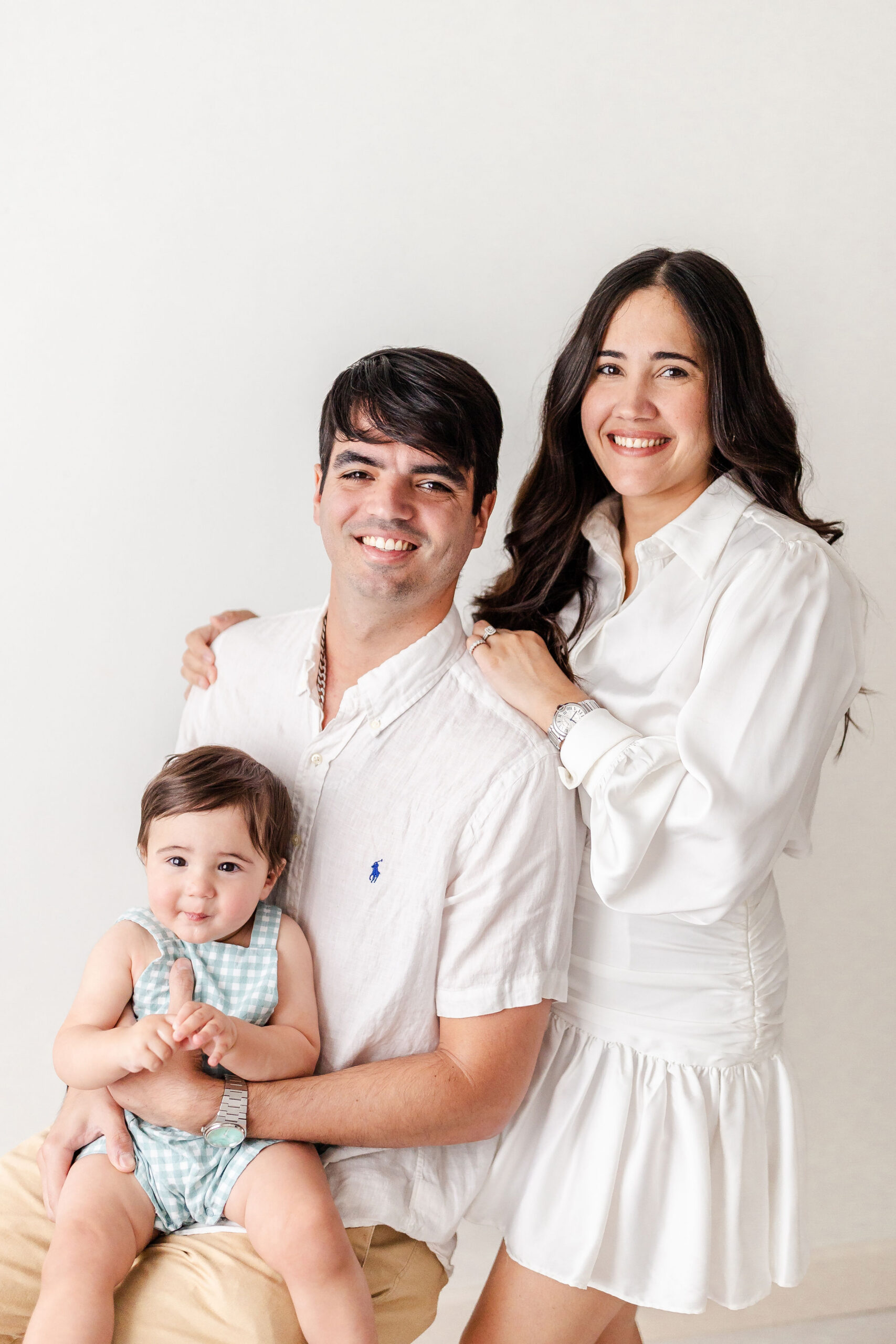 A happy mom and dad in a studio in white with baby boy in blue suspenders in dad's lap after exploring state of kid