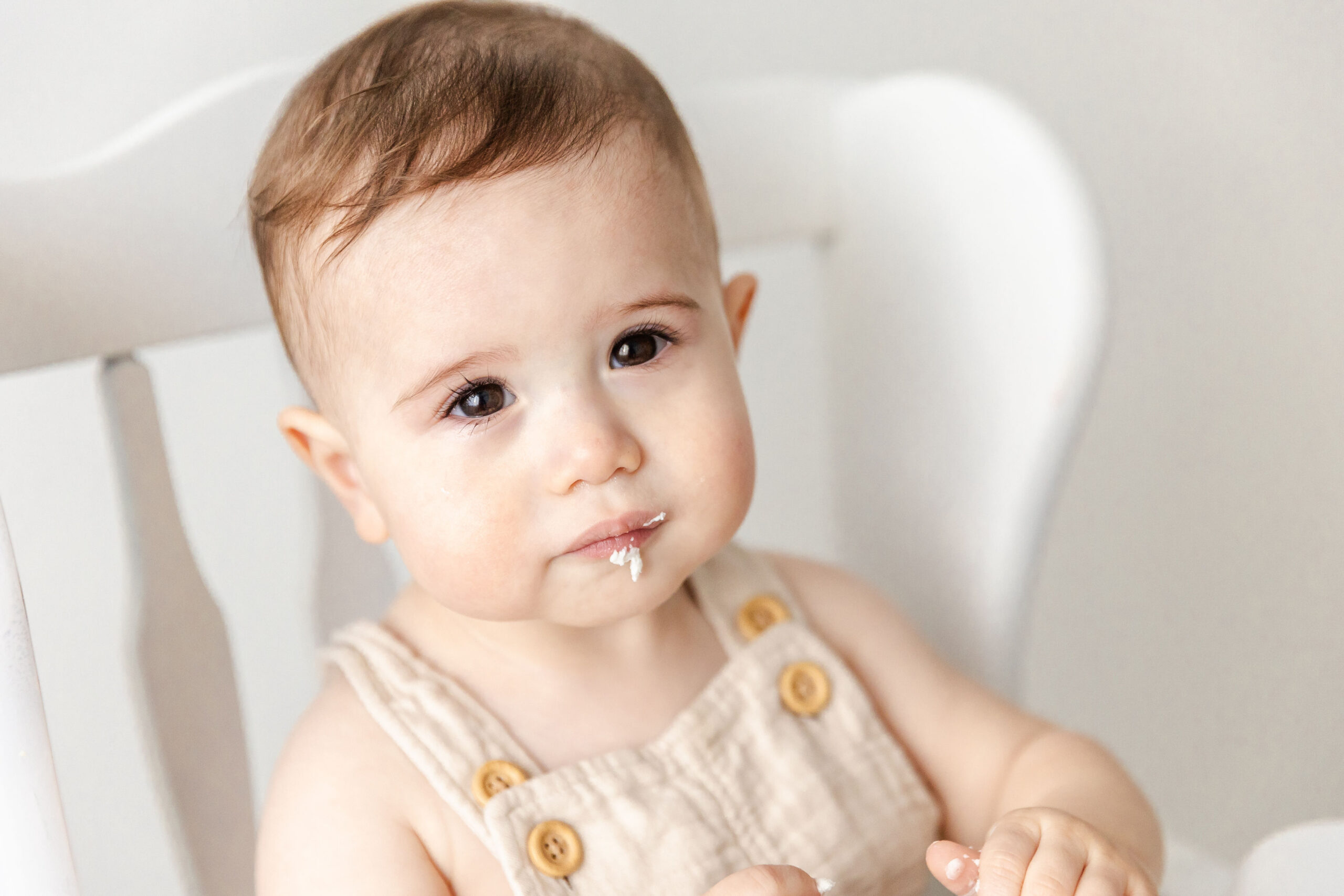 A closeup of a baby boy in cream suspenders with dark eyes and icing on his lips